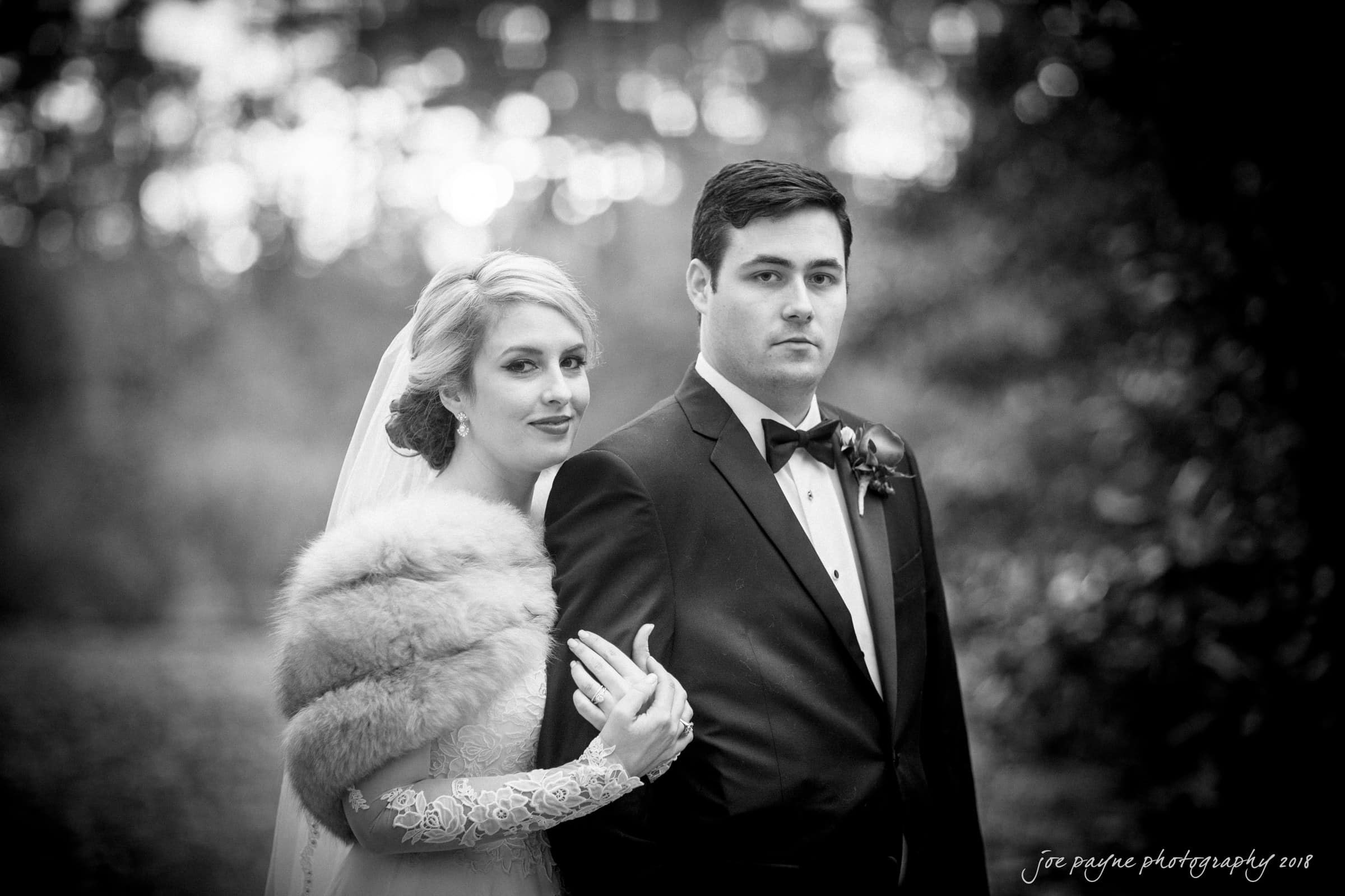 A bride and groom pose outdoors in a black and white photo. The bride is wearing a veil and fur stole, while the groom is in a tuxedo. They stand closely, with trees blurred in the background.