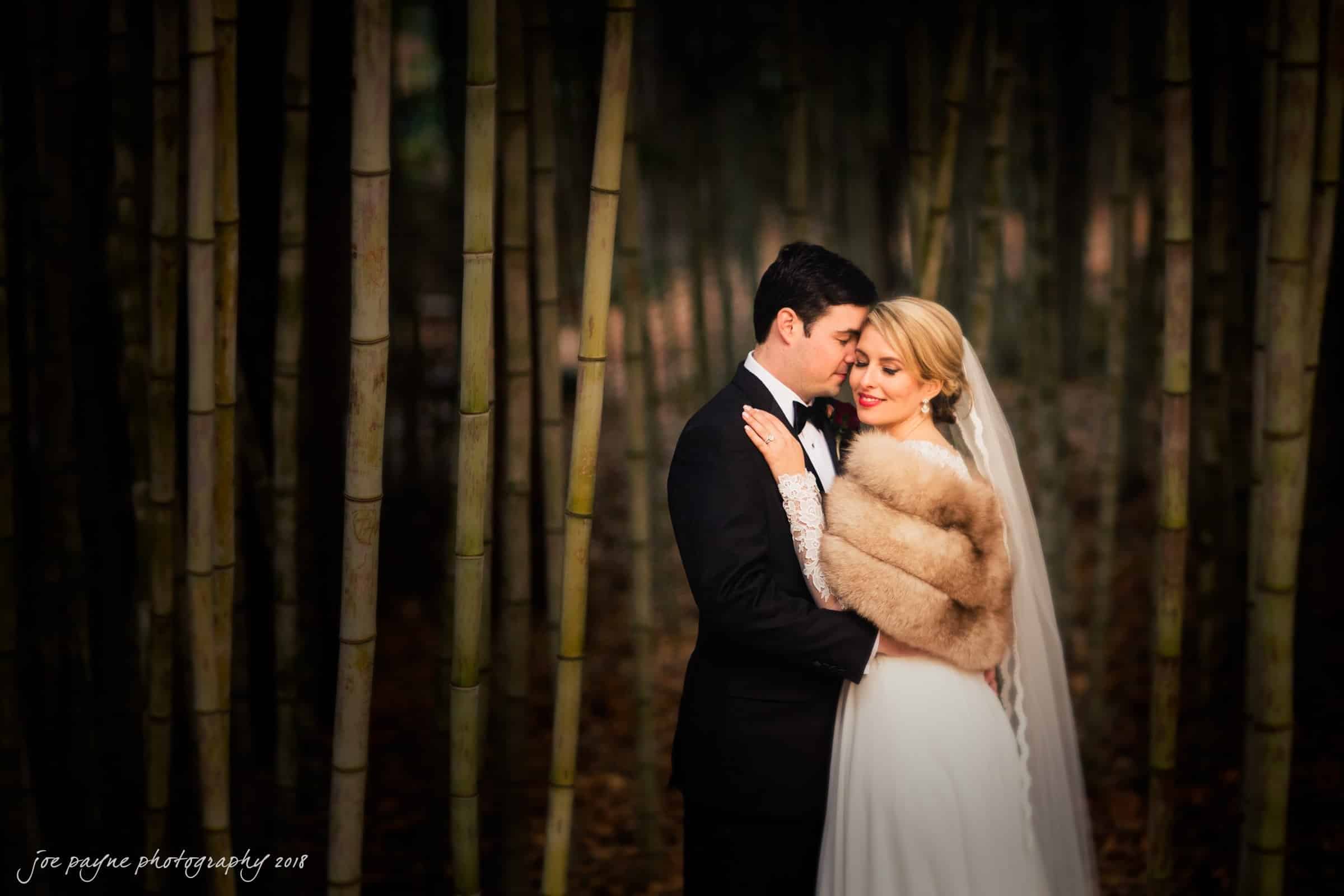 A bride and groom embrace in a forest of tall bamboo. The bride wears a long white dress and a fur stole, while the groom is in a black suit. The scene is romantic and serene, with soft lighting creating a warm atmosphere.