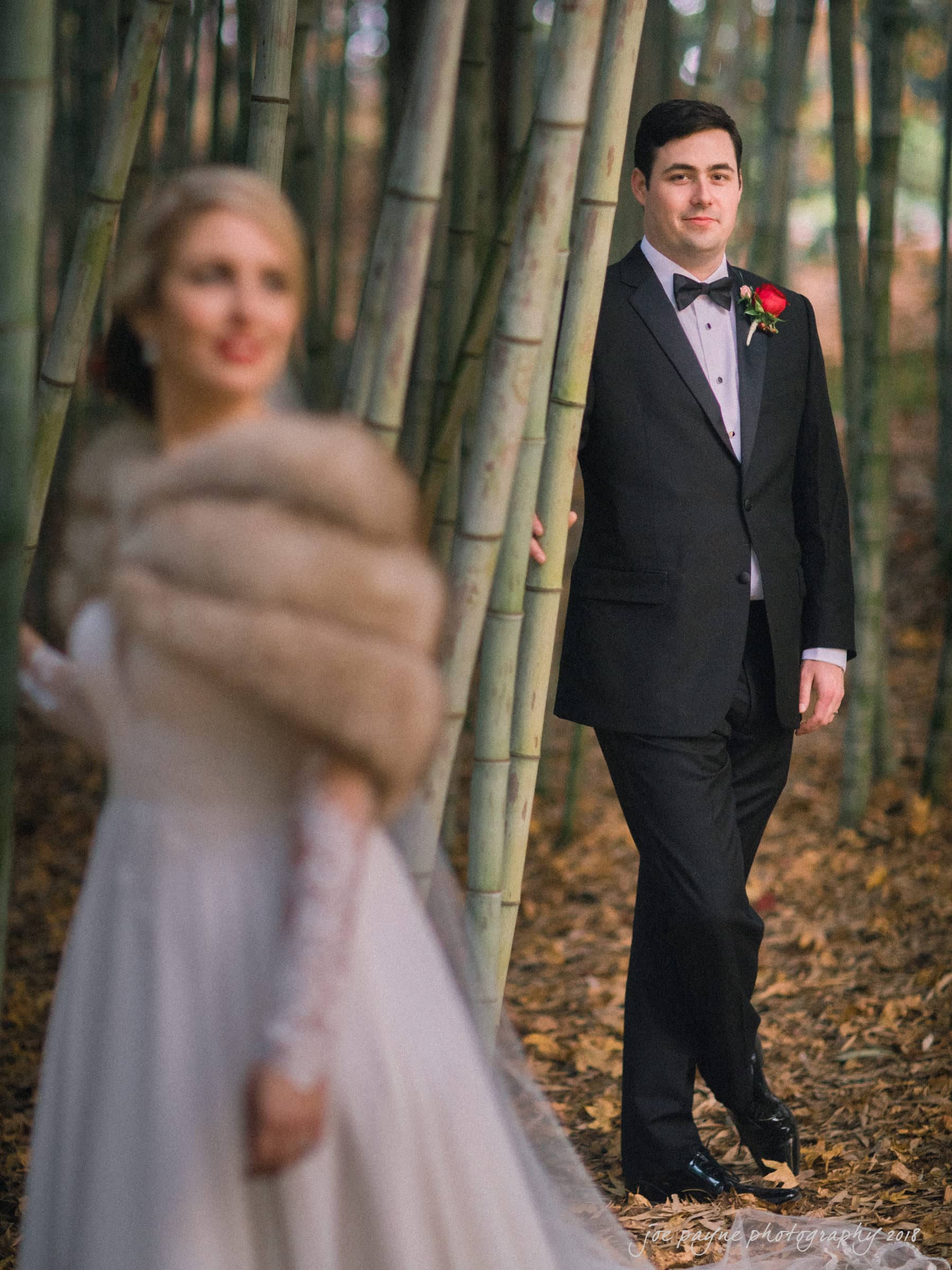 A groom, in a black tuxedo with a red rose boutonniere, stands among bamboo. In the foreground, slightly blurred, is a bride in a white dress with long lace sleeves and a fur stole. The setting is natural with fallen leaves.