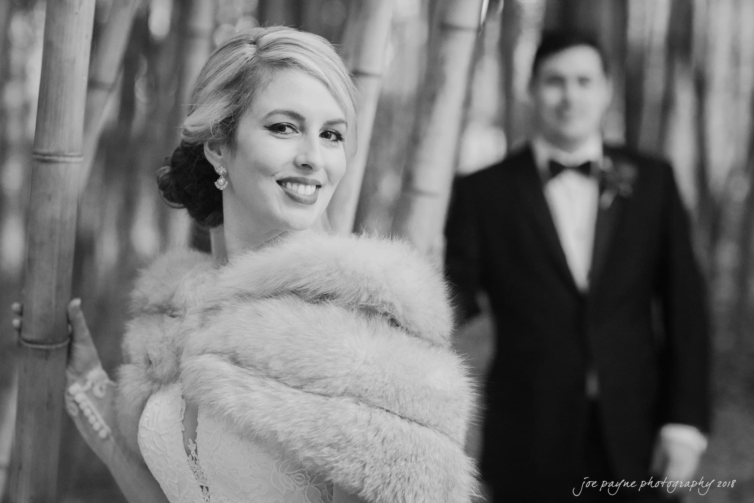 A joyful bride smiles at the camera, wearing a lace dress and a fur stole. In the blurred background, a groom in a tuxedo stands among tall bamboo stalks. The black and white image captures a romantic, elegant outdoor setting.