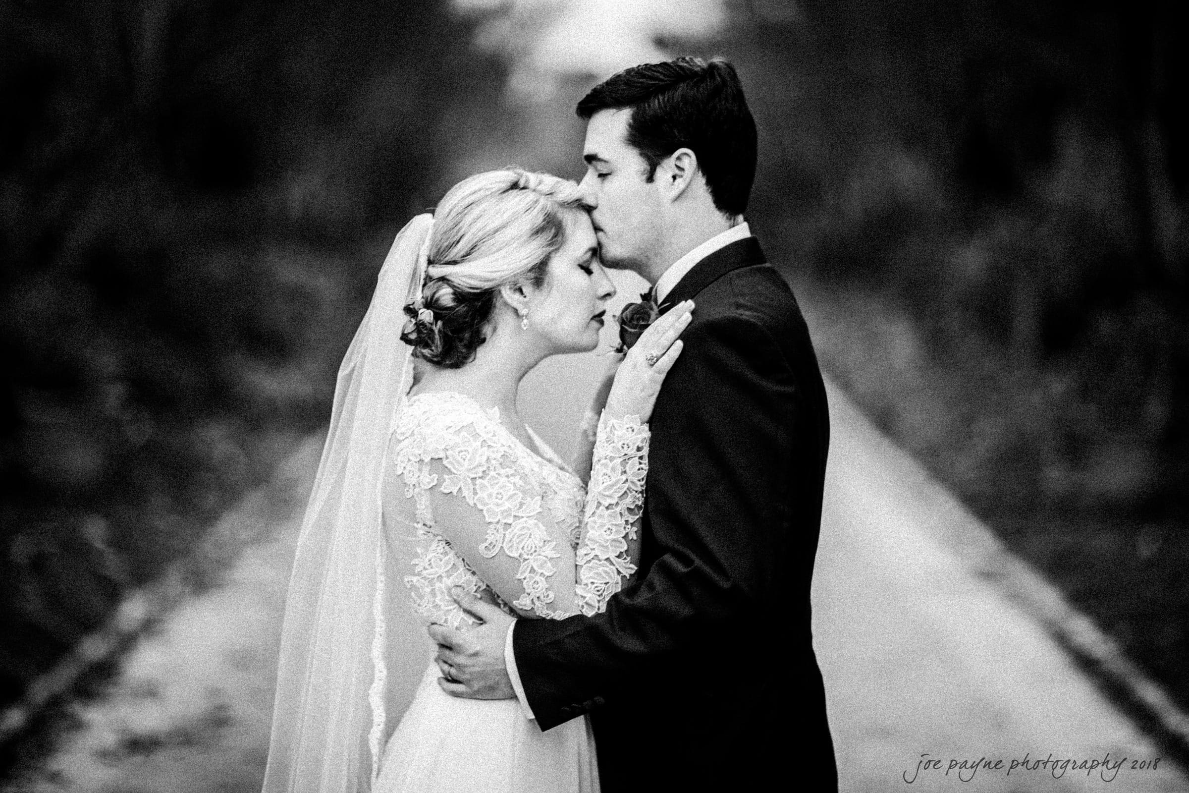 A bride and groom stand close on a path surrounded by trees. The groom gently kisses the brides forehead. She wears a lace wedding dress and veil; he is in a dark suit. The scene captures an intimate moment.