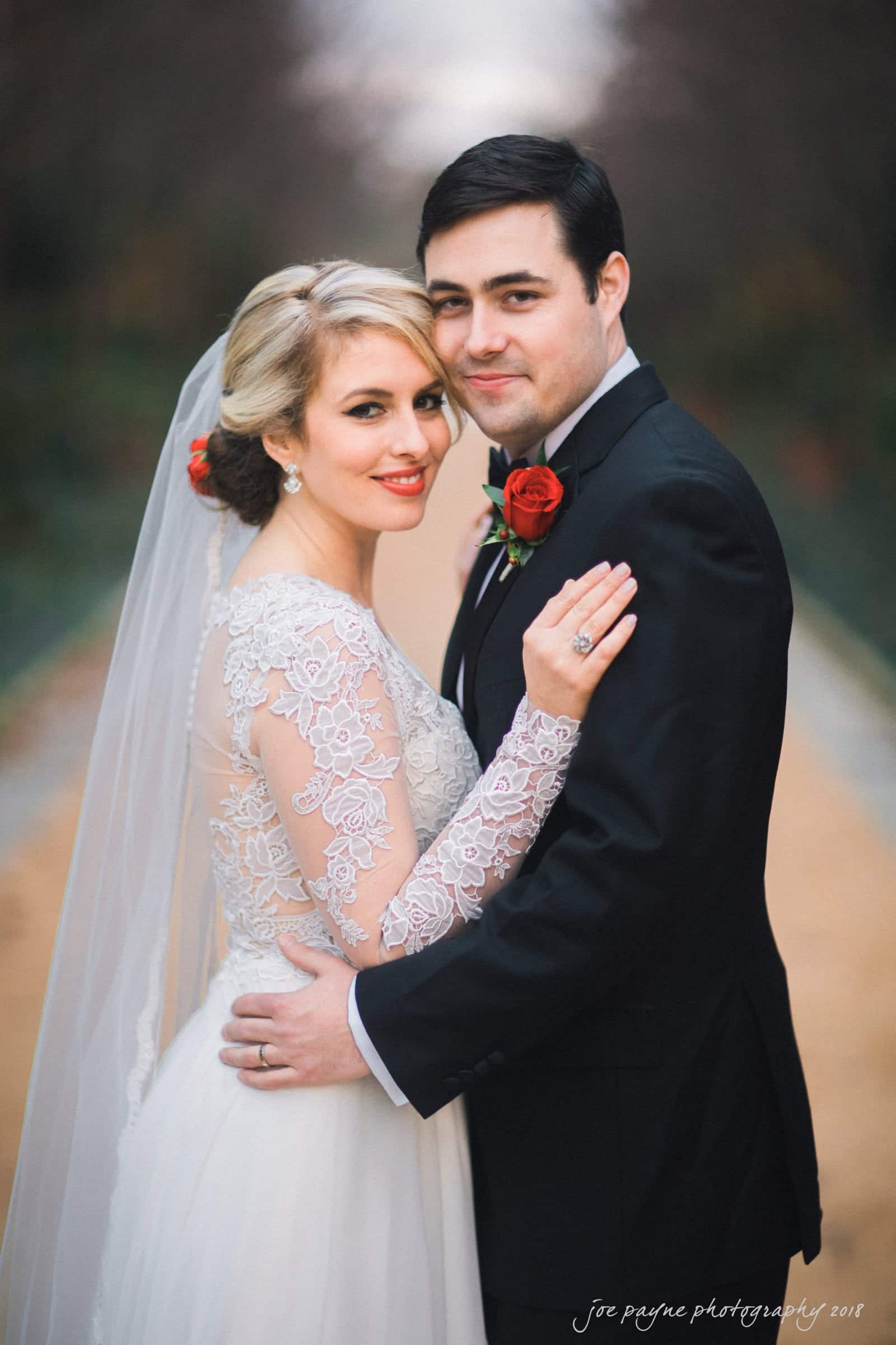 A bride and groom pose outdoors on a path lined with greenery. The bride wears a lace gown and veil, while the groom is in a black suit with a red rose boutonniere. They smile and embrace on their wedding day.