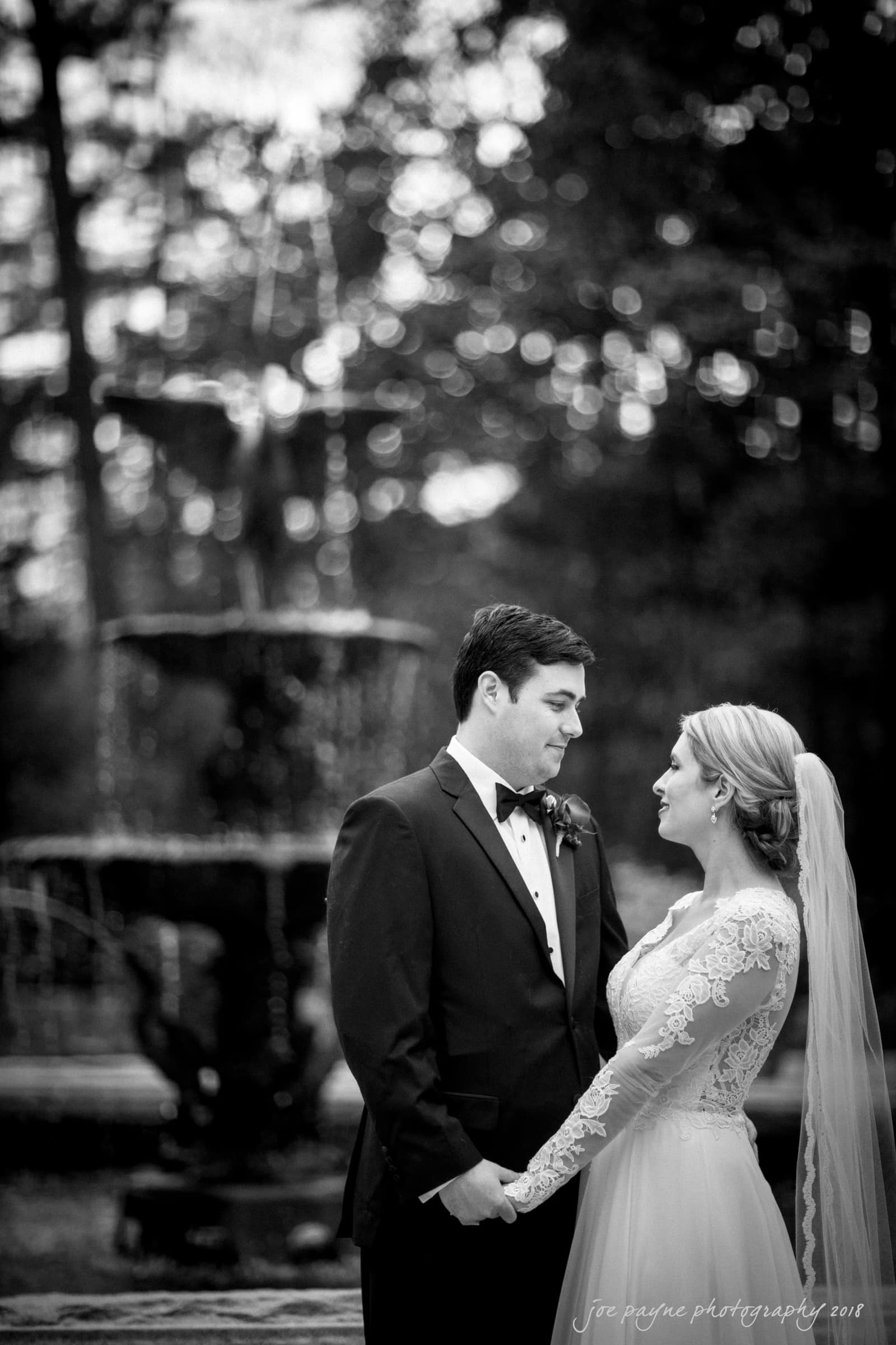 A black and white photo of a bride and groom holding hands and gazing into each others eyes. The bride wears a long-sleeved lace gown and veil, the groom is in a dark suit. A blurred fountain is visible in the background.