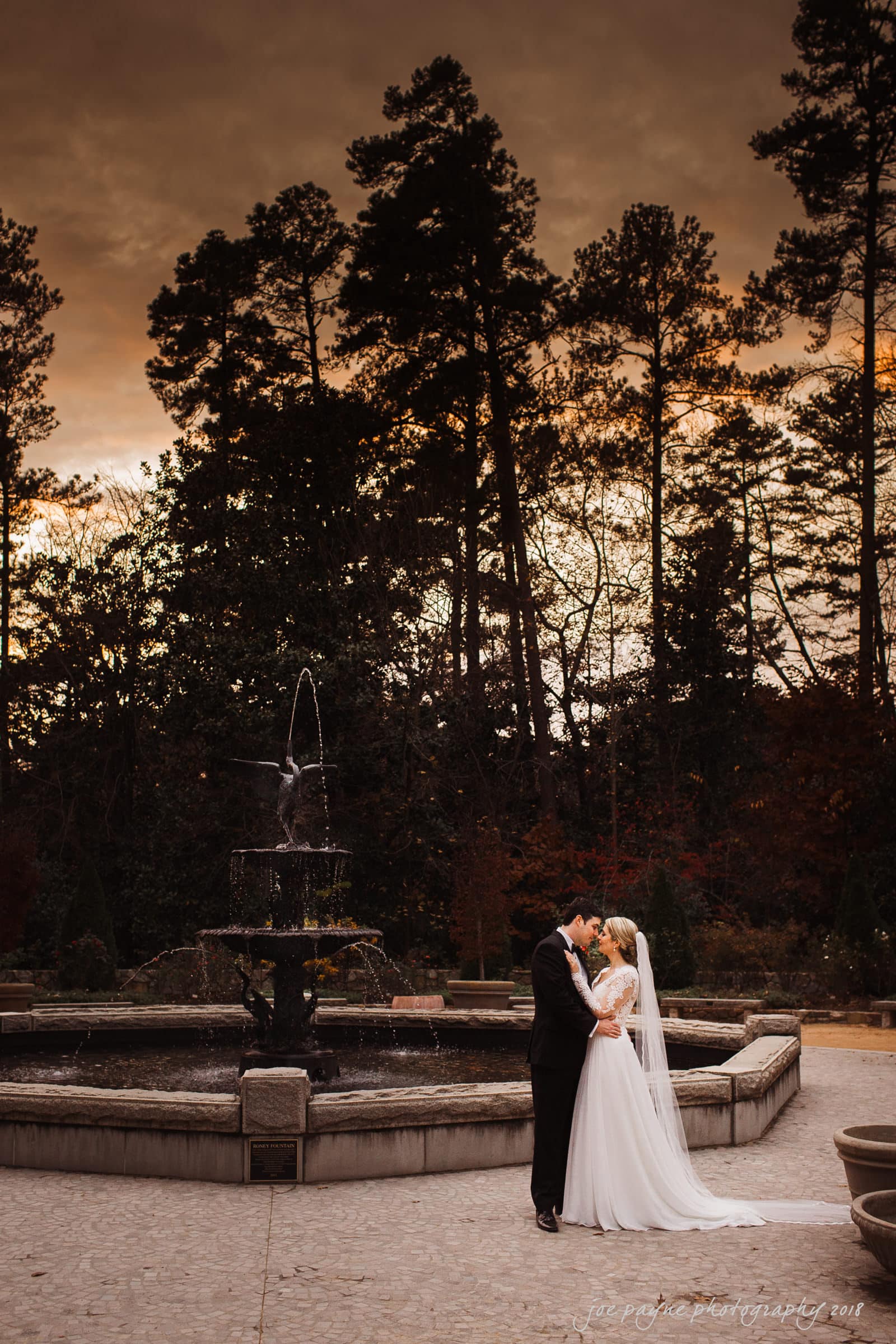 A bride and groom share a kiss beside a fountain at sunset. The couple is embracing, surrounded by tall trees and a dramatic sky. The setting evokes a romantic and serene atmosphere.
