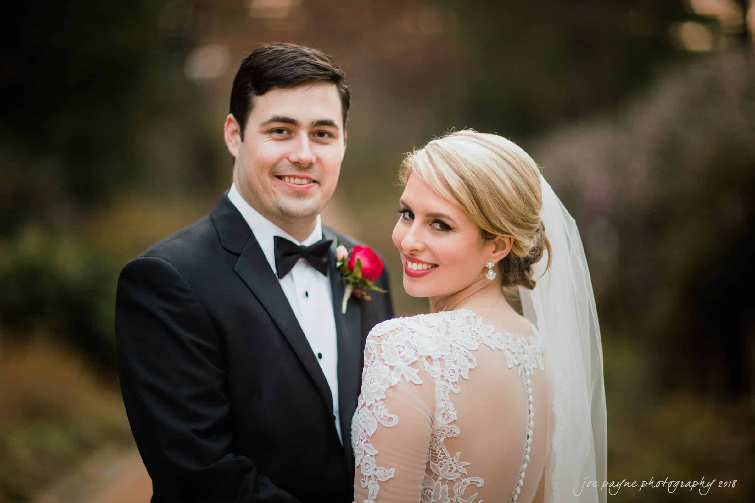 A bride and groom smile warmly at the camera. The bride wears an elegant lace gown with a veil, and the groom is in a black tuxedo with a red rose boutonniere. They stand outdoors with a blurred natural background.