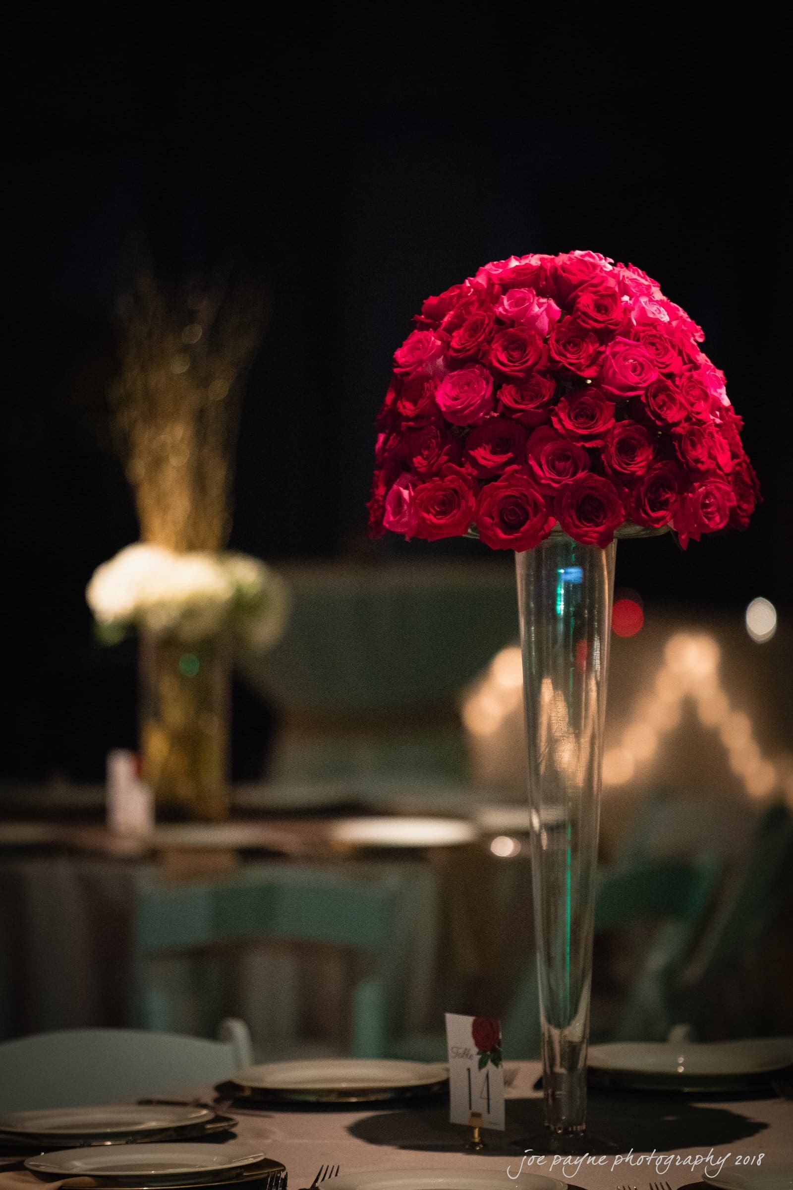 A tall, elegant centerpiece with vibrant red roses in a clear vase sits on a table set with plates and silverware. Soft lighting highlights the centerpiece, with blurred background elements and another floral arrangement visible.