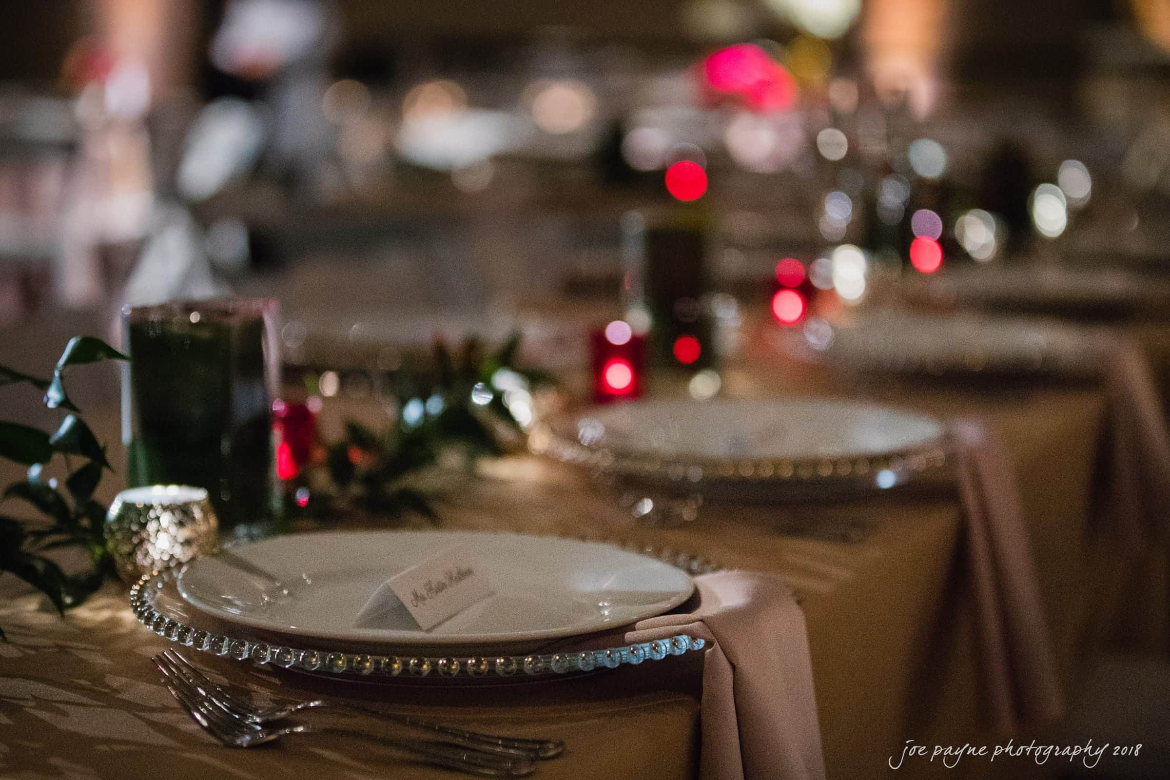 A dimly lit elegant dining table set up with sparkling charger plates and name cards. The table is adorned with green foliage and small red candles, creating a warm and intimate ambiance.