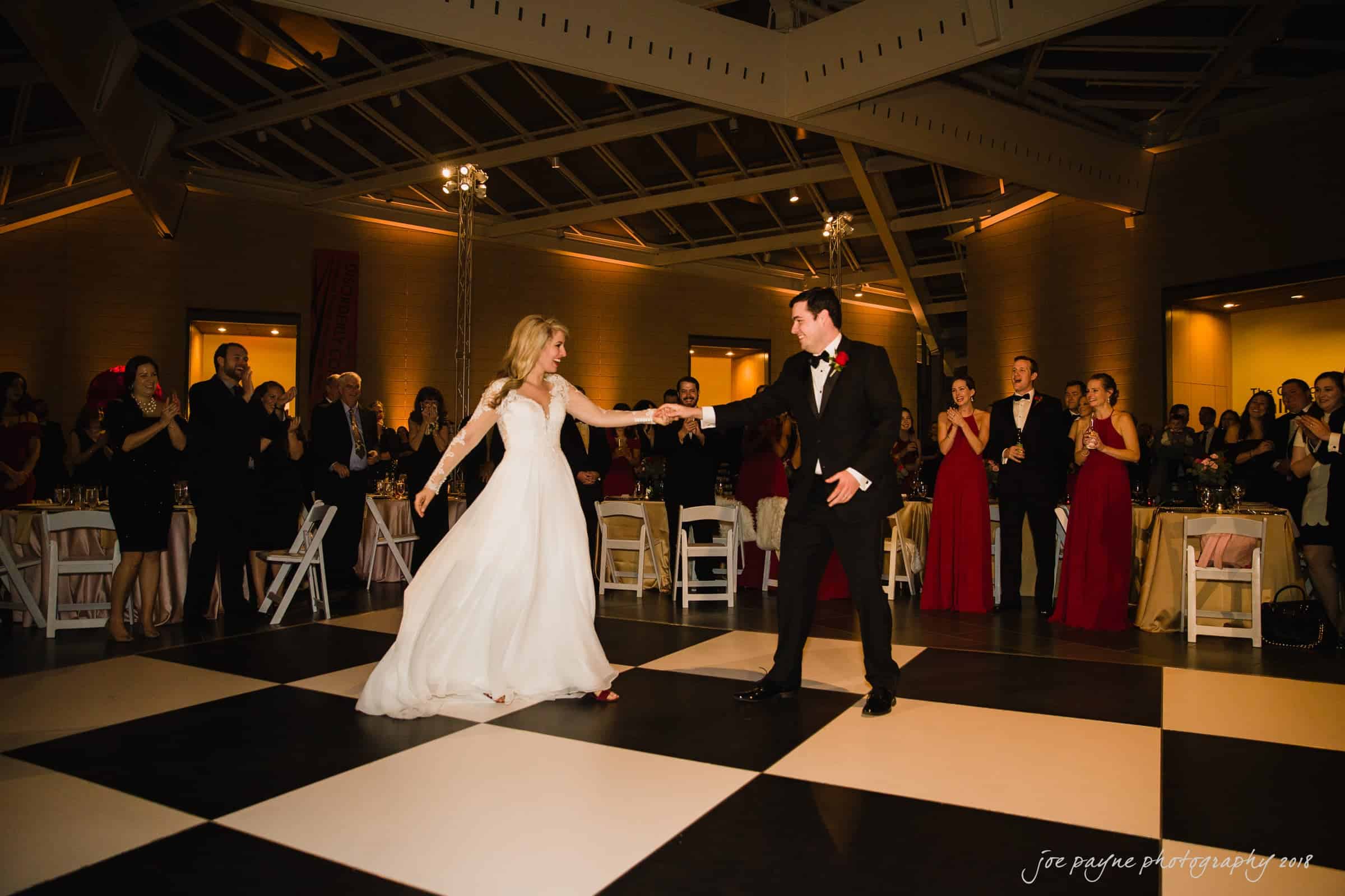 A couple dances elegantly on a black and white checkered floor at a formal event. The woman wears a long white dress, and the man wears a black suit with a red boutonniere. Guests in elegant attire watch and clap in the background.