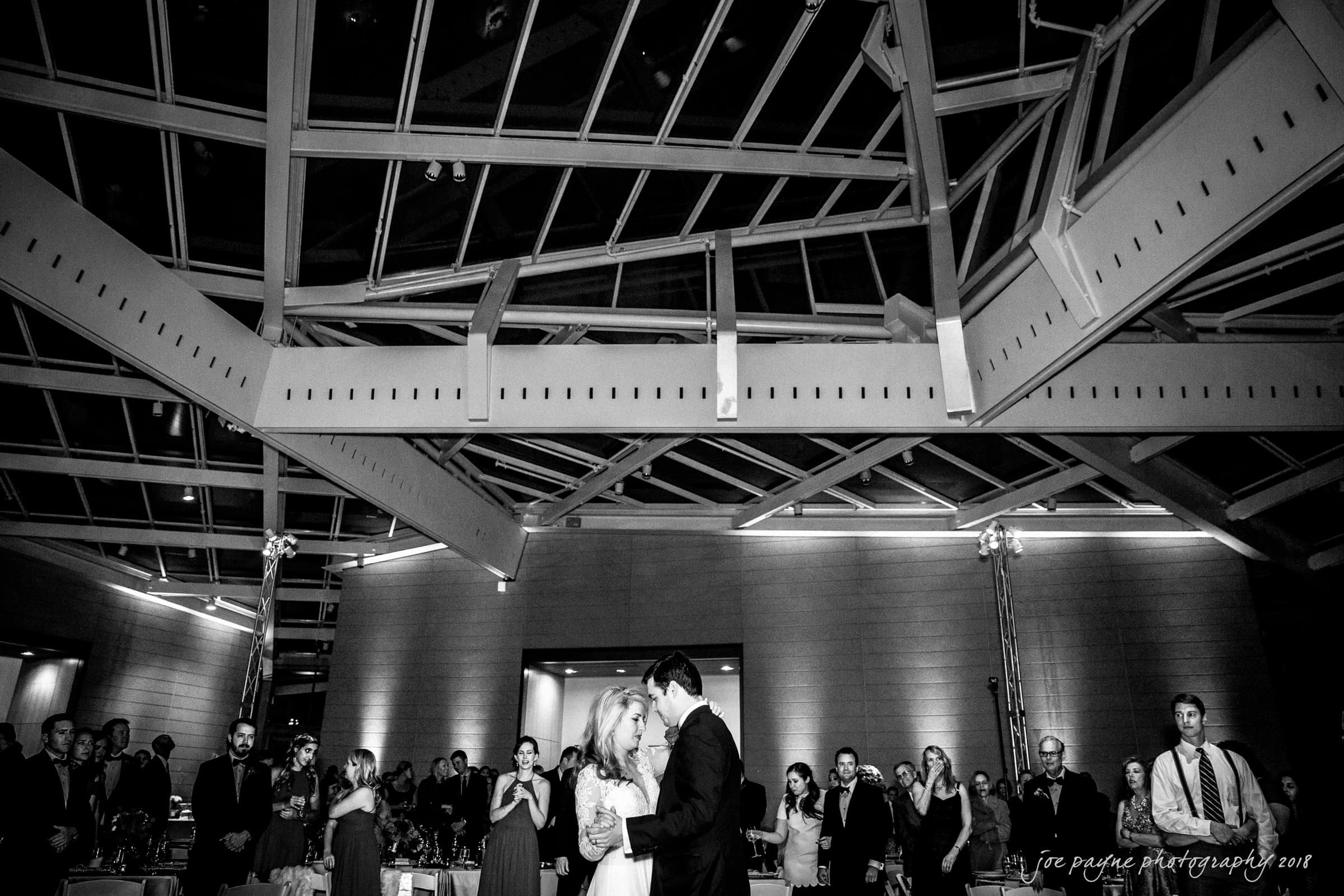 A bride and groom share their first dance in a modern, glass-roofed venue. Guests stand around, watching the couple with smiles. The room is elegantly lit, highlighting the romantic atmosphere.