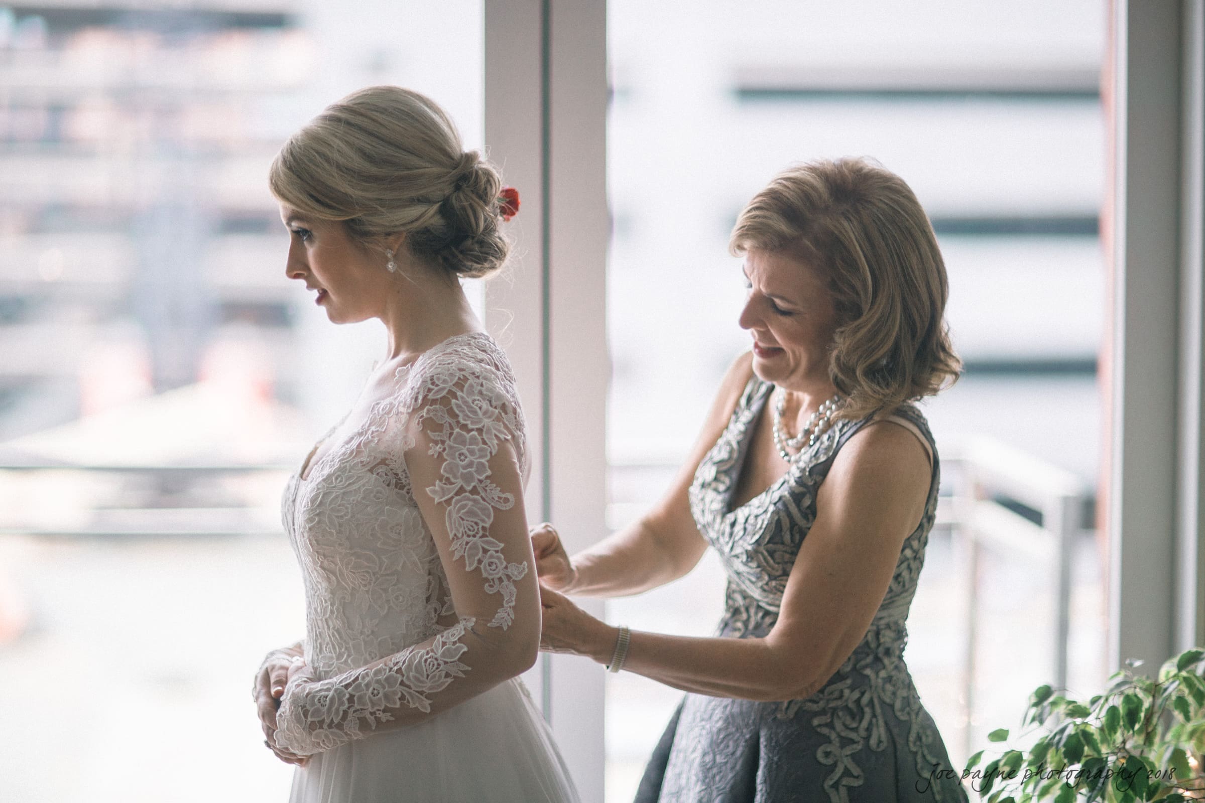 A bride in a lace wedding dress stands by a window, while another woman in a patterned dress helps adjust the back of her gown. The scene is softly lit, with an urban background visible through the window.