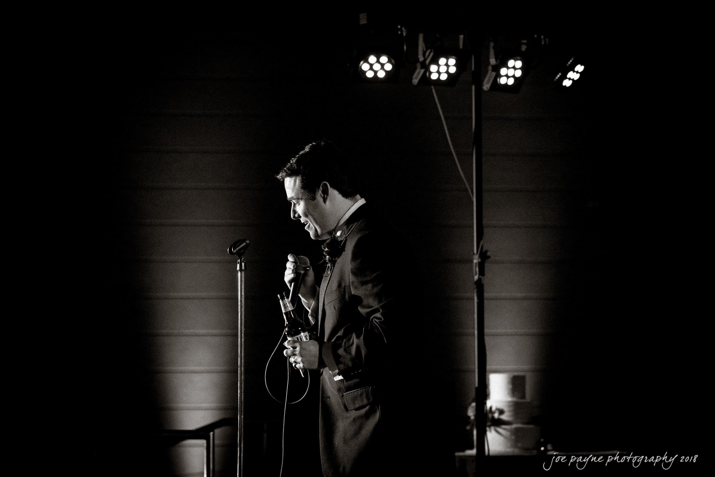 A man in a suit stands in profile holding a microphone in his left hand, under stage lights against a dark background. The mood is dramatic, with a spotlight on him.