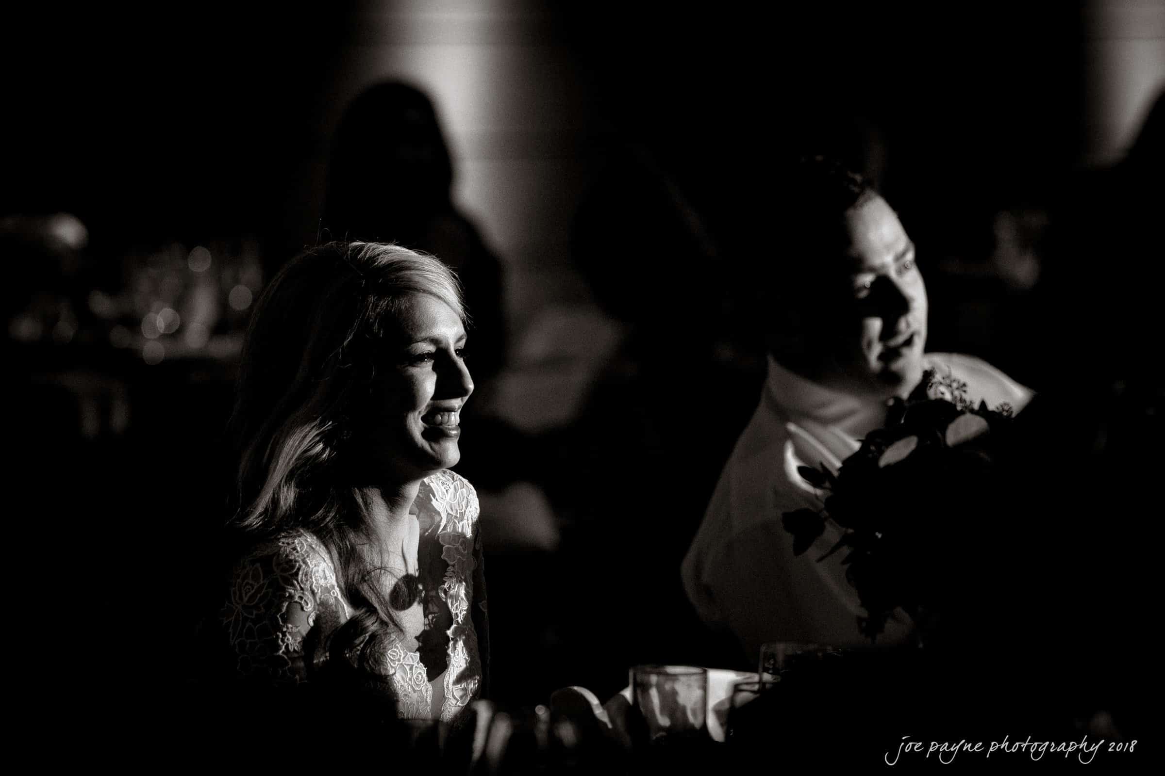 A black-and-white photo of a smiling couple sitting together. The woman is on the left, wearing a light-colored lace dress, while the man on the right is wearing a suit, holding a bouquet. The background is softly blurred.