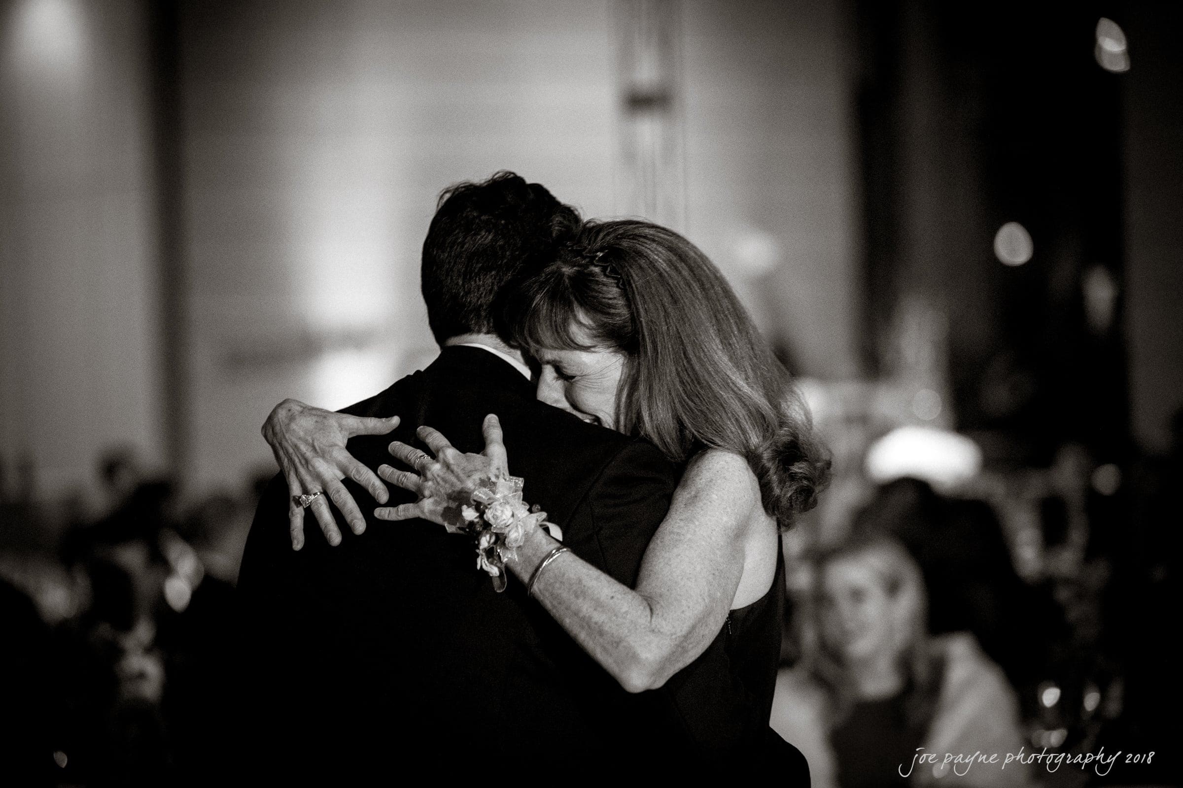 A couple is dancing closely at what appears to be a formal event. The woman, wearing a wrist corsage, embraces the man with her eyes closed. The background is softly blurred, creating an intimate atmosphere.