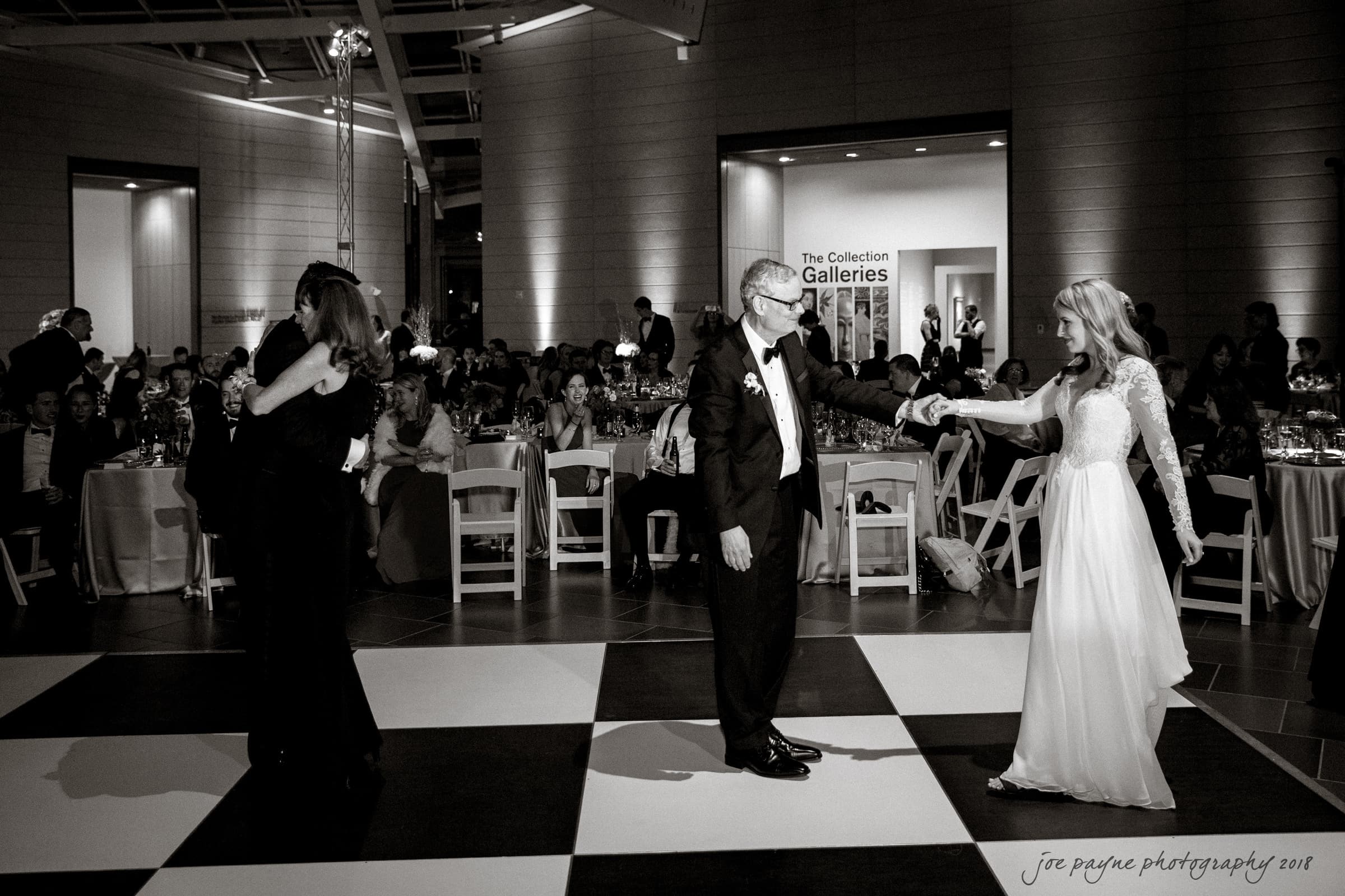 A black and white photo of a wedding reception. Couples are dancing on a large checkered floor. In the foreground, a bride in a flowing dress dances with an older man in a tuxedo. Guests sit at tables in the background.