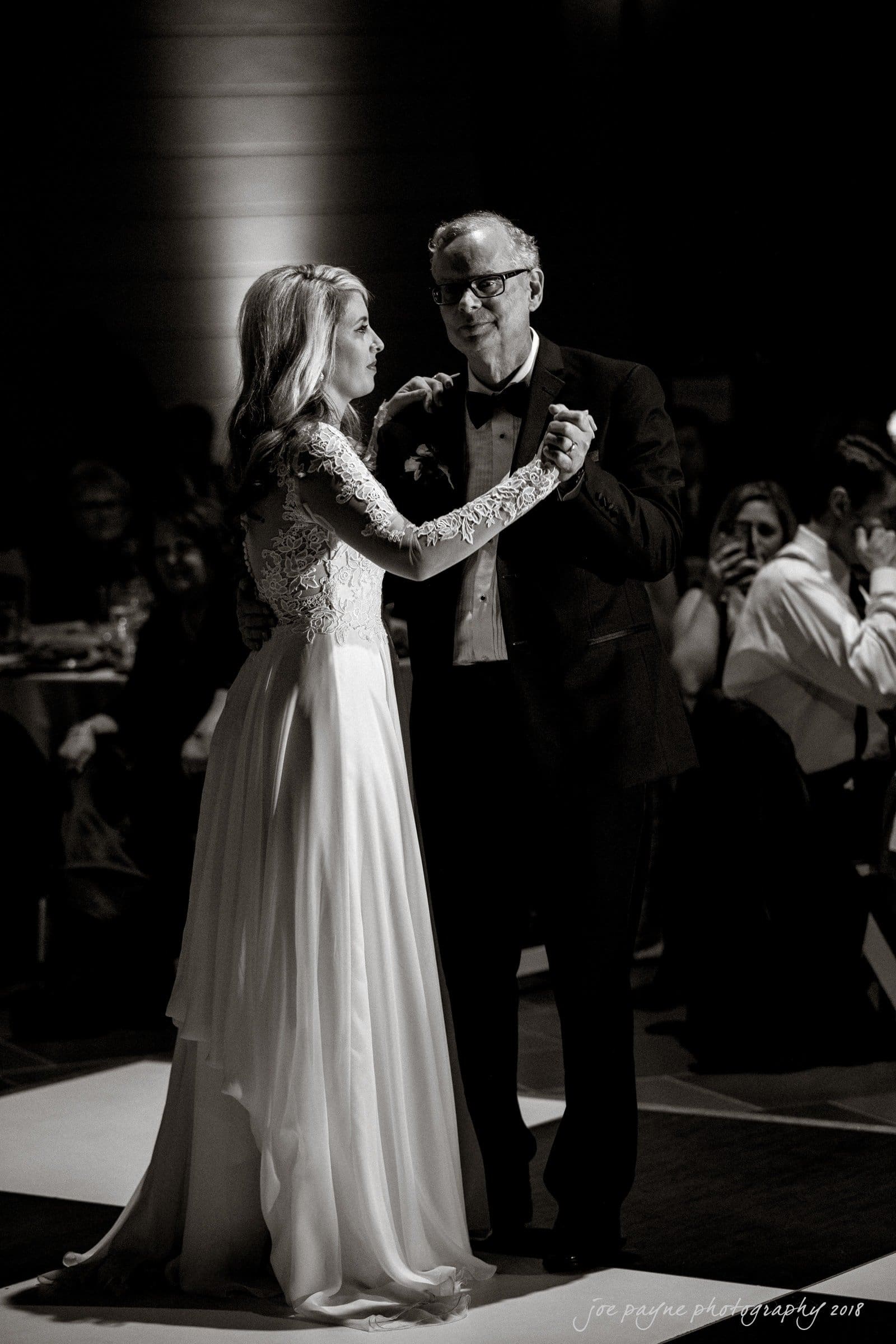 A bride and an older man, both elegantly dressed, dance together on a dimly lit dance floor. The woman wears a long, flowing gown with lace sleeves. Guests watch them in the background, seated at tables. The mood is intimate and joyous.