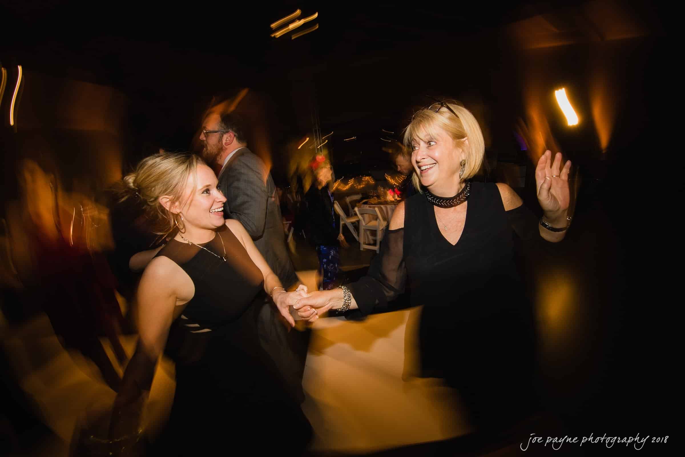 Two women in black dresses dance joyfully at a party. The motion blur adds a dynamic feel, with warm lighting creating a lively ambiance. Other guests appear in the background.