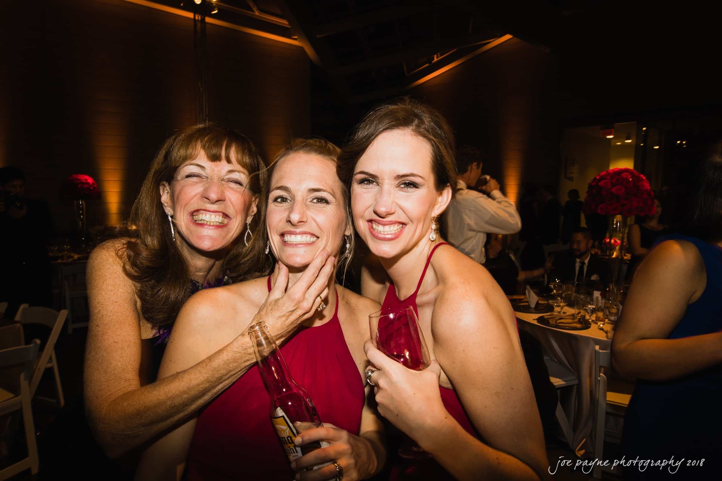 Three women smiling and posing for a photo at an indoor event. Two are in matching red dresses, holding glasses, while the third woman stands behind them, laughing. The background shows warmly lit tables and a red flower arrangement.