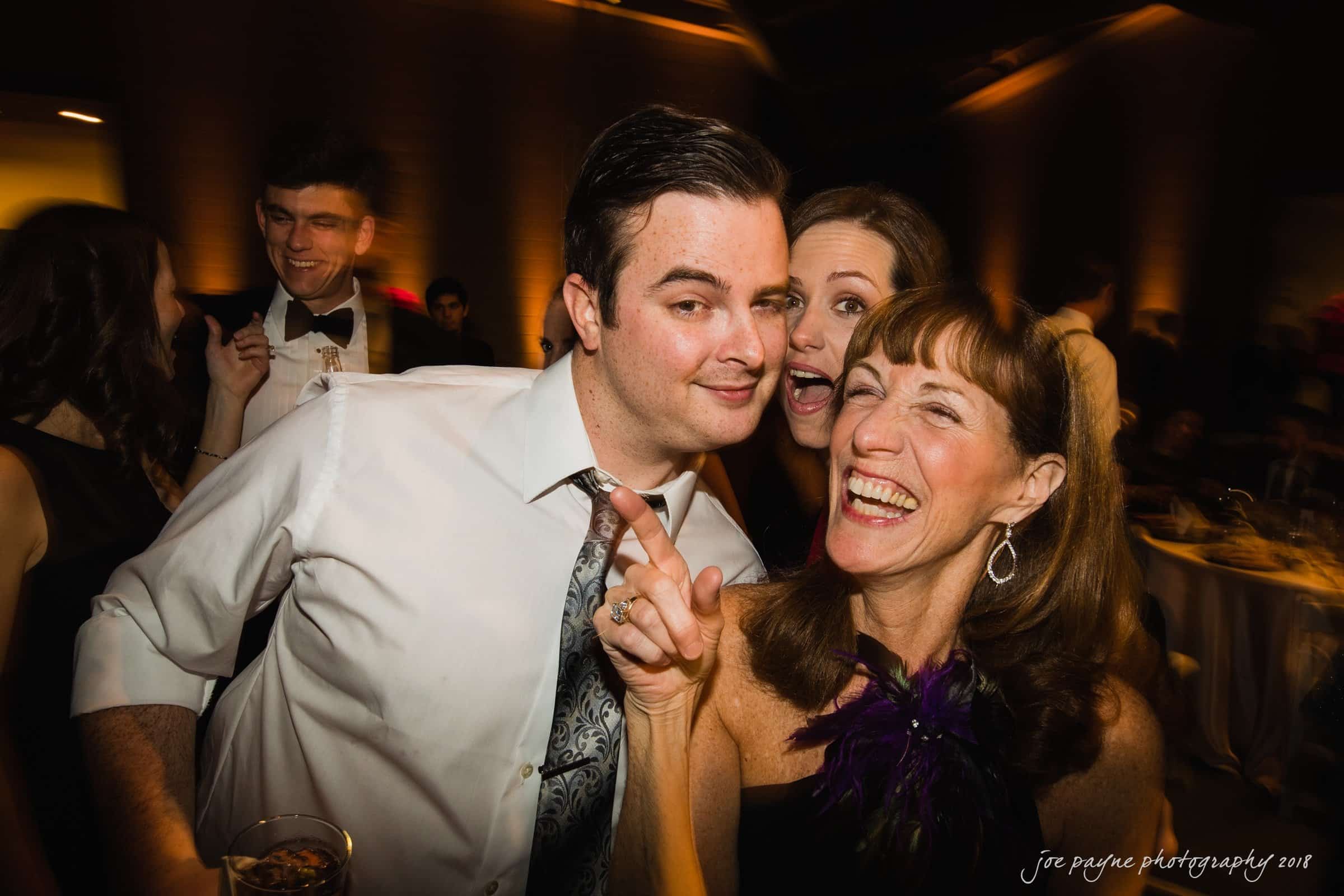 A group of people enjoying a festive event. A woman in a purple and black dress is smiling and pointing, while a man in a white shirt stands beside her. Others are socializing in the background. The atmosphere is lively and celebratory.