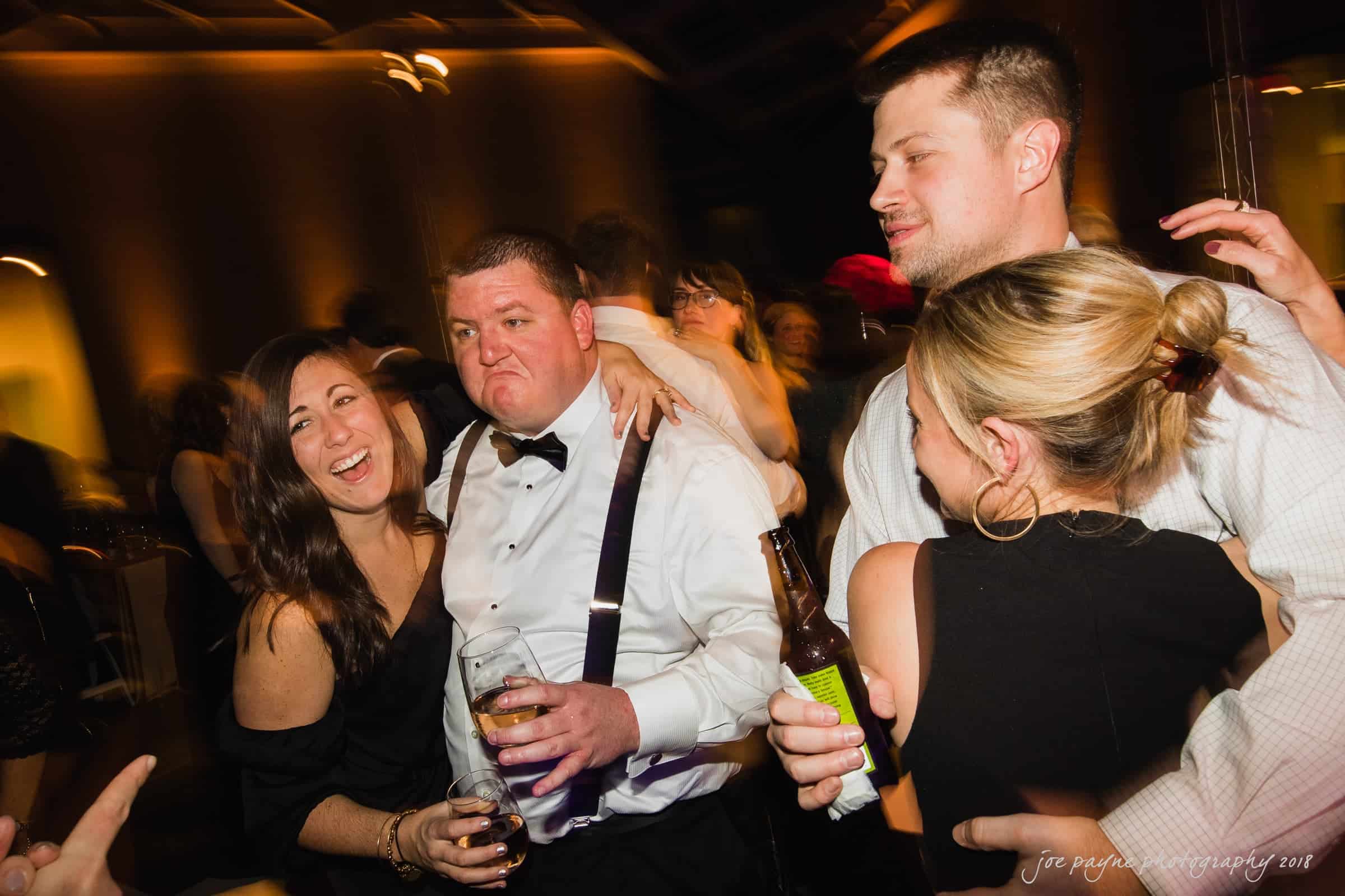 A group of people enjoying a night out at a party. A man wearing a white shirt and suspenders is in the center, surrounded by smiling friends. They hold drinks and appear to be having a lively time.