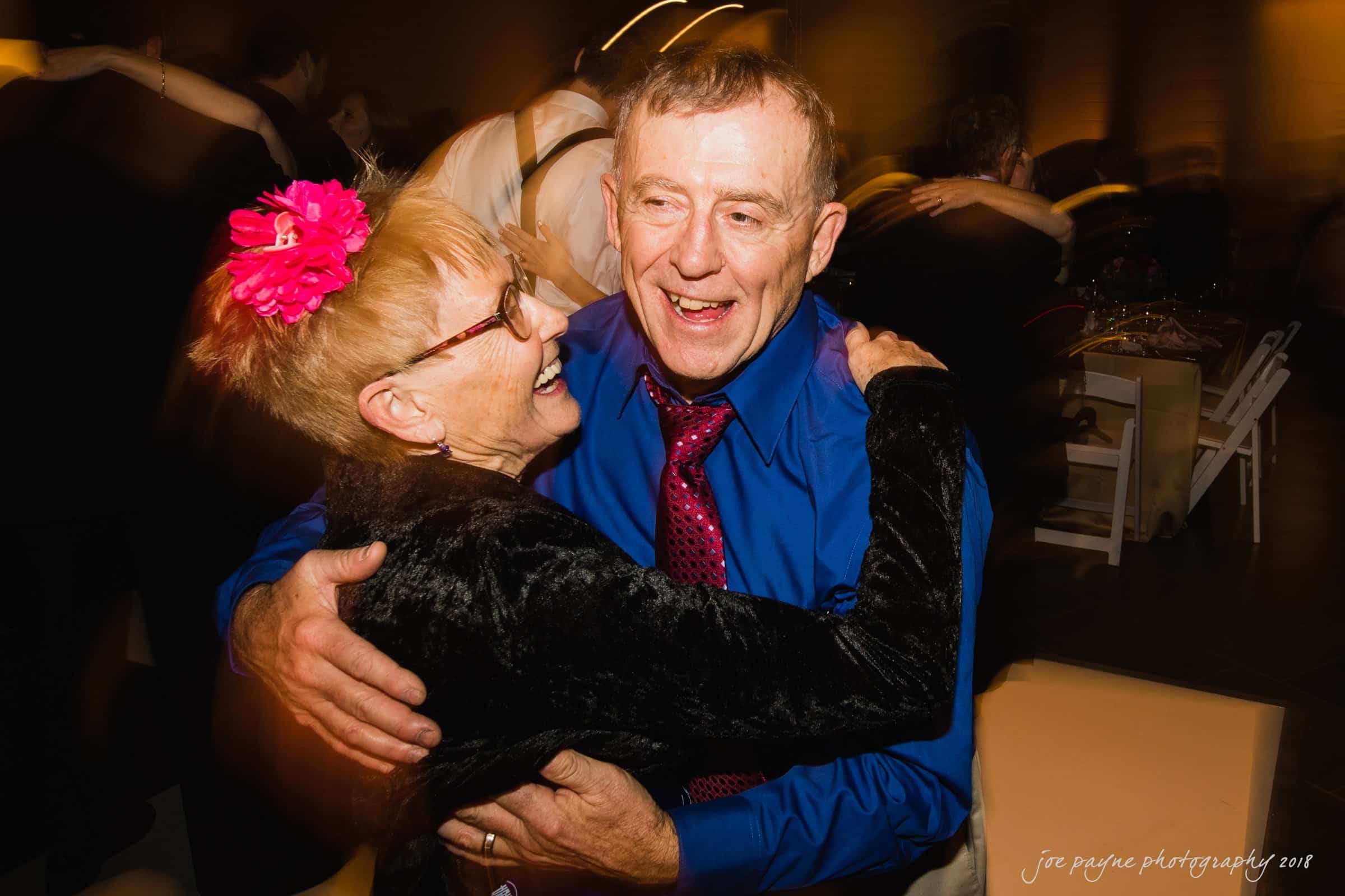 An elderly couple joyfully dance together at a lively event. The woman wears a bright pink flower in her hair and glasses, while the man sports a blue shirt and patterned tie. Both are smiling and embracing each other on the dance floor.