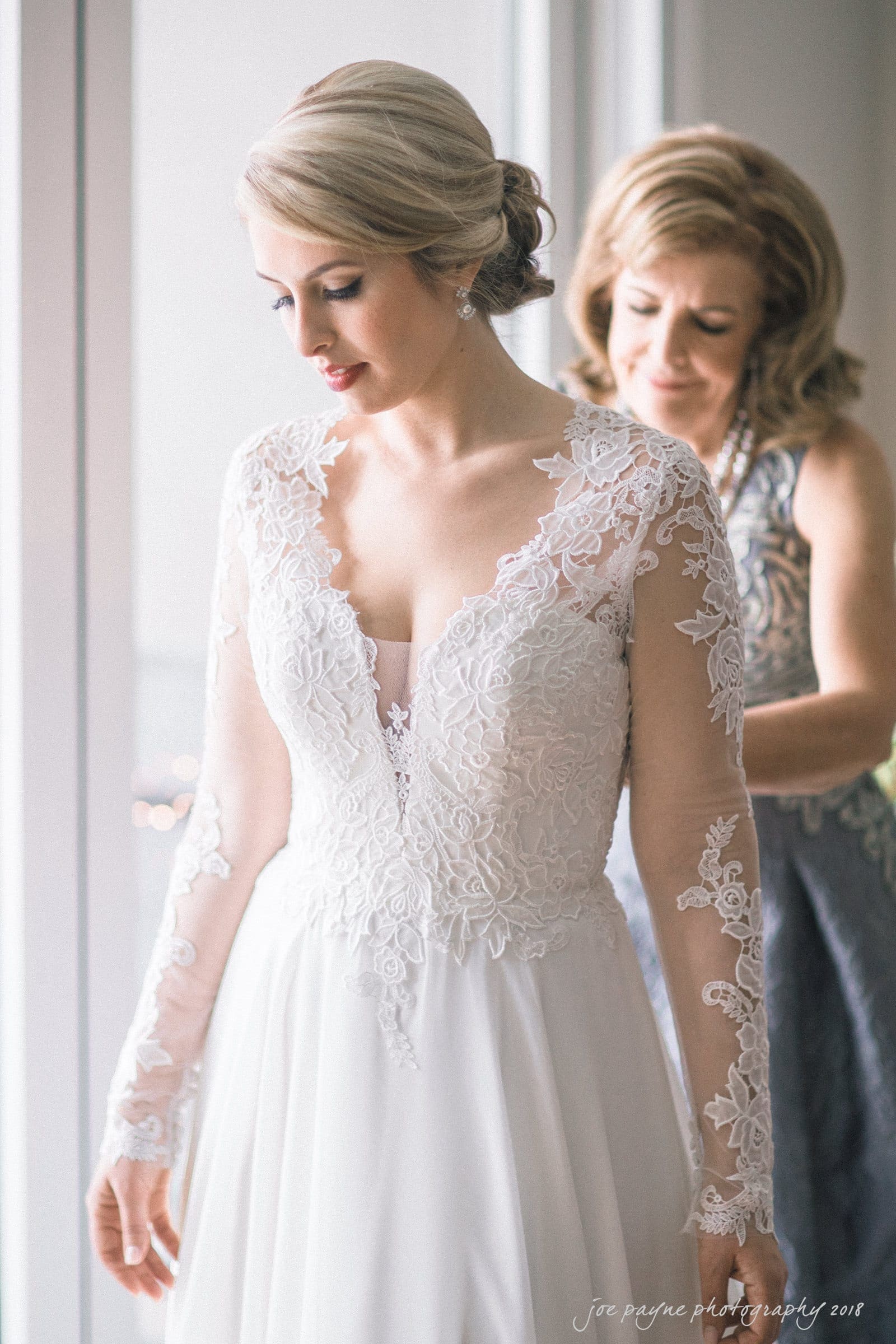 A bride in a lace wedding dress stands by a window while an older woman assists with adjustments. The dress has intricate floral details and long sleeves. The scene is serene and well-lit, with soft natural light.
