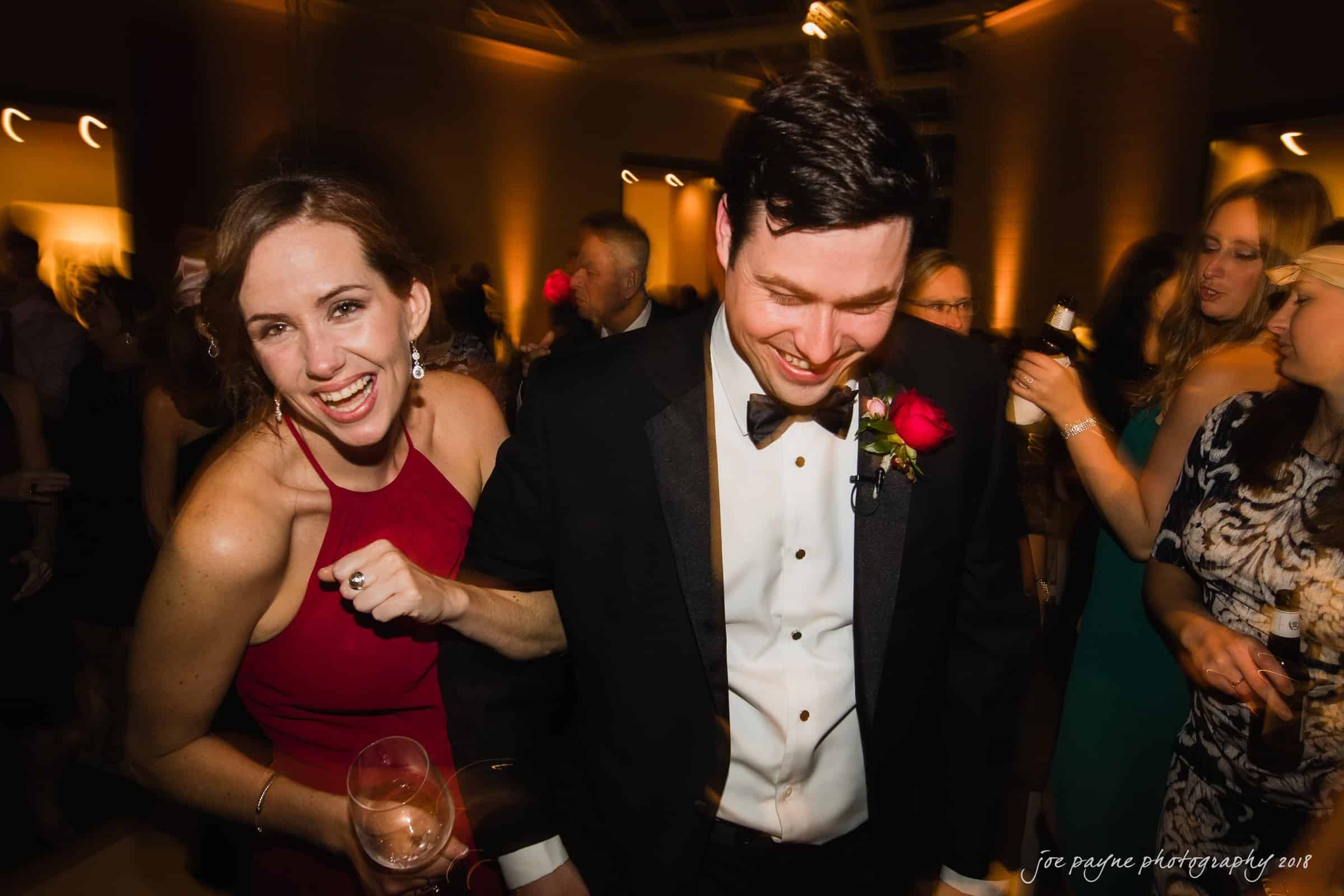 A smiling woman in a red dress and a man in a tuxedo with a red rose are joyfully walking together at a lively indoor event. People are mingling and holding drinks in the softly lit background.