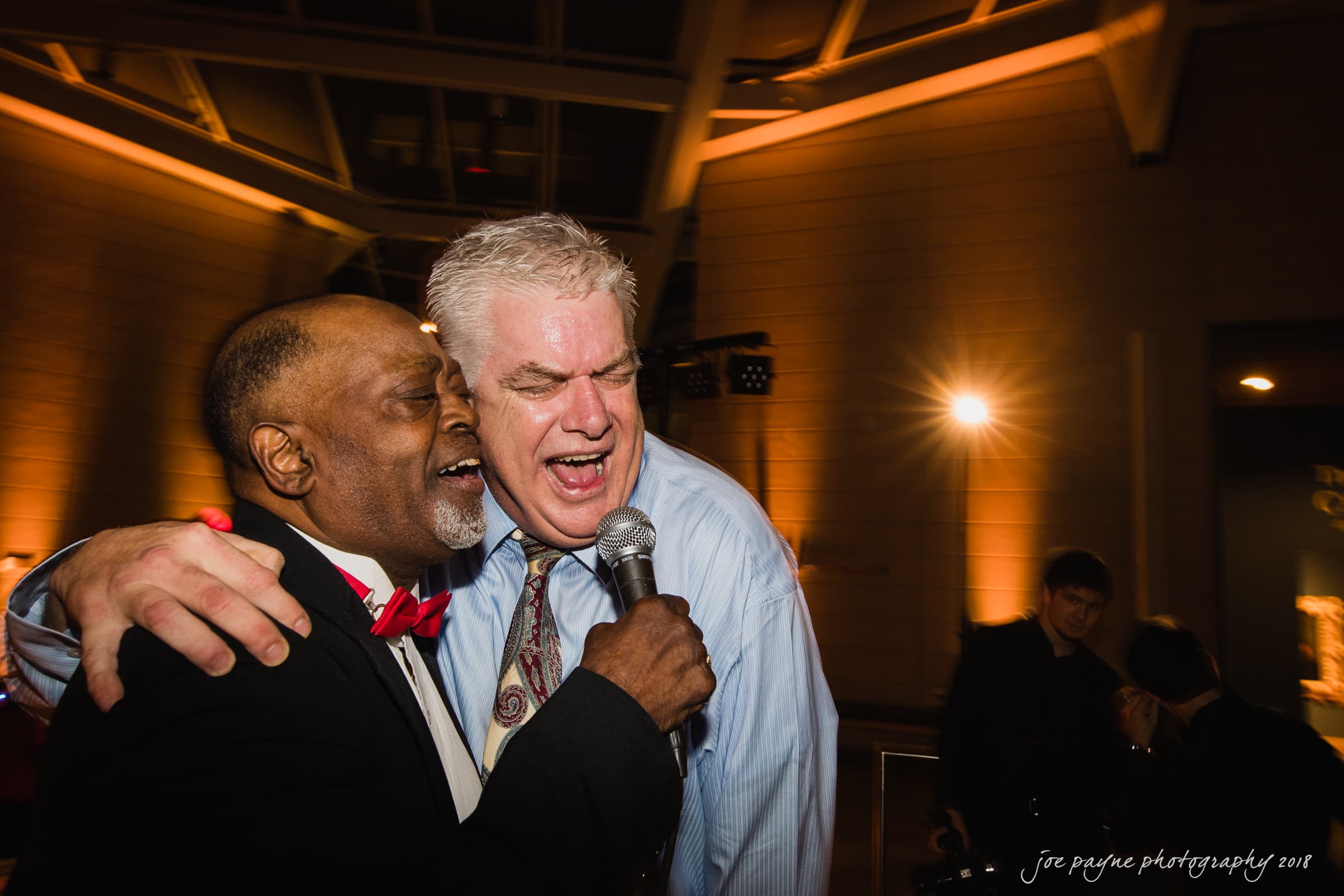 Two men singing energetically into a microphone at an indoor event. One is wearing a suit with a red bow tie, and the other is in a dress shirt and tie. Theyre surrounded by ambient lighting, creating a lively atmosphere.