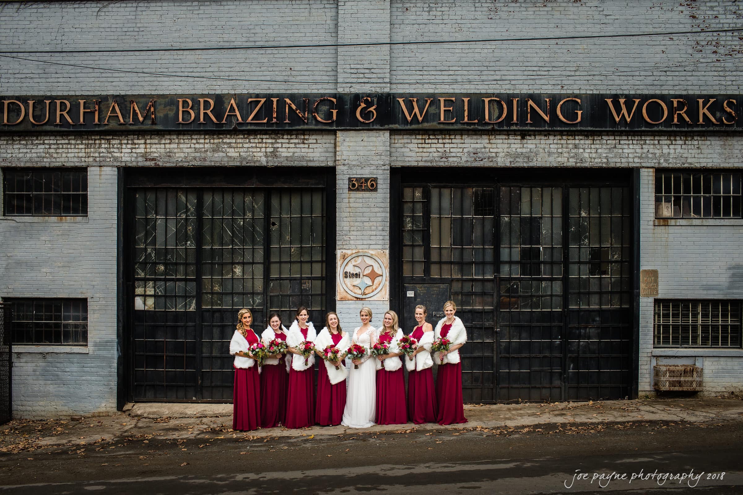 A group of bridesmaids in burgundy dresses and white shawls stand with the bride in front of a rustic, industrial building with large windows and the sign Durham Brazing & Welding Works. The women are holding floral bouquets.