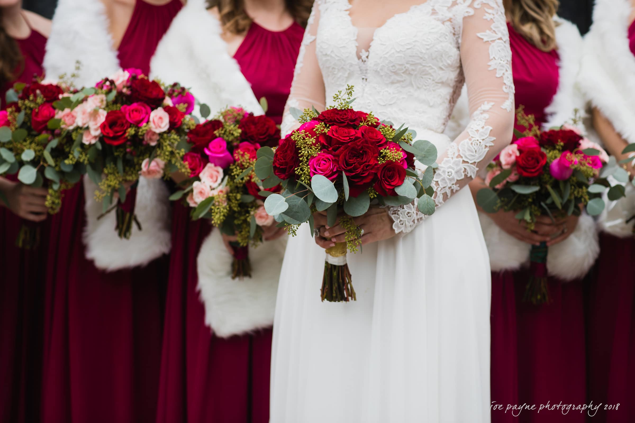 A bride in a lace wedding dress holds a bouquet of red roses and greenery. She is surrounded by bridesmaids holding similar bouquets, dressed in burgundy dresses with white fur shawls.