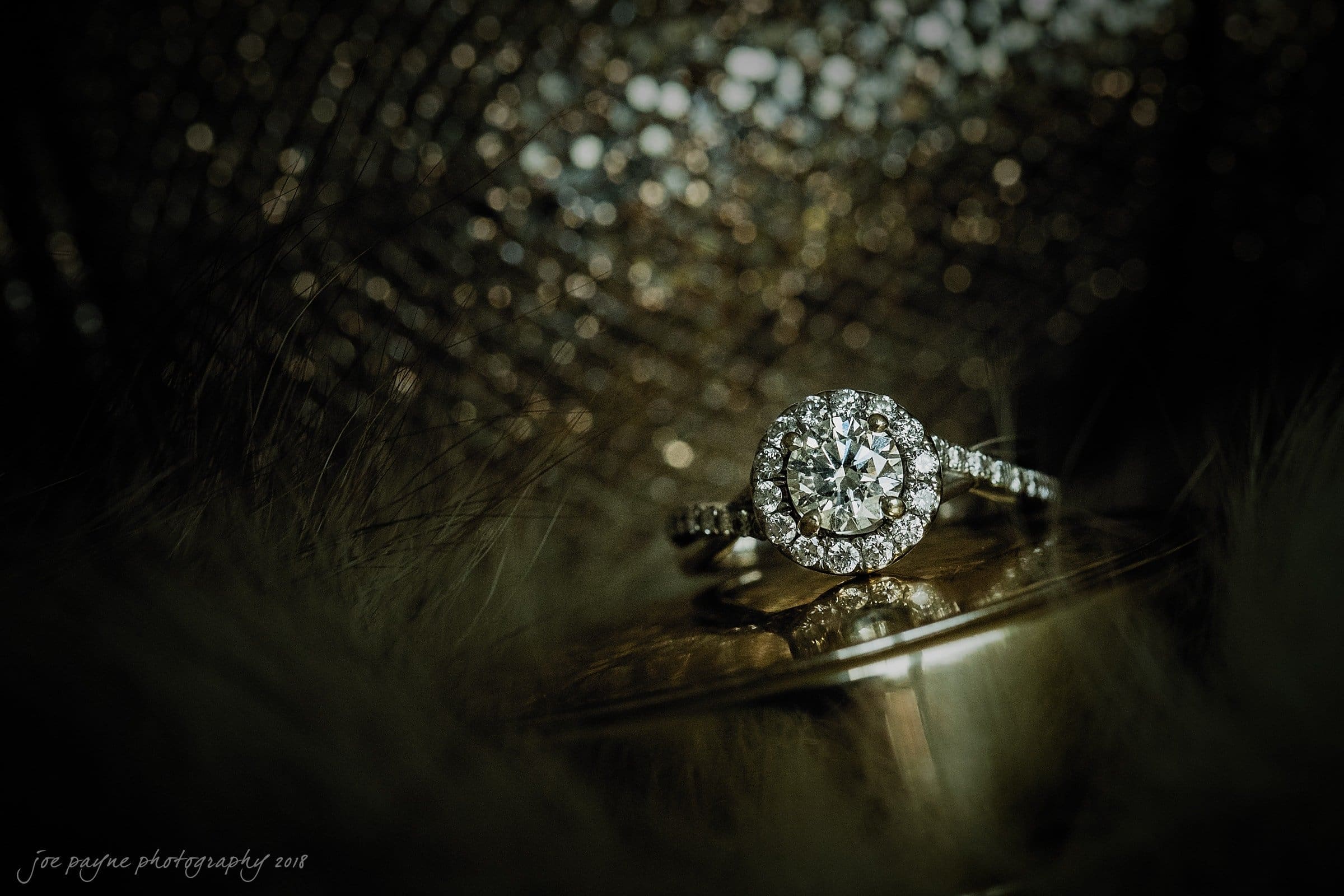 A sparkling diamond ring with a halo setting rests on a metallic surface, surrounded by a softly blurred background. The rings intricate details glitter under the light, creating a luxurious and elegant atmosphere.