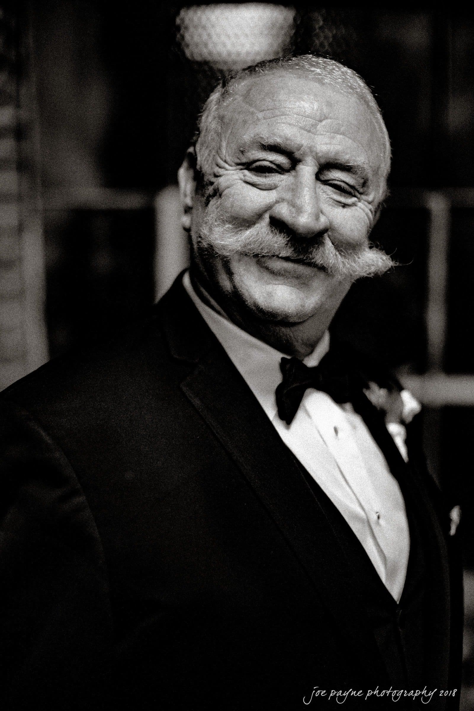 Elderly man in a tuxedo with a prominent mustache smiles warmly. The setting is dimly lit, creating a classic and elegant atmosphere.