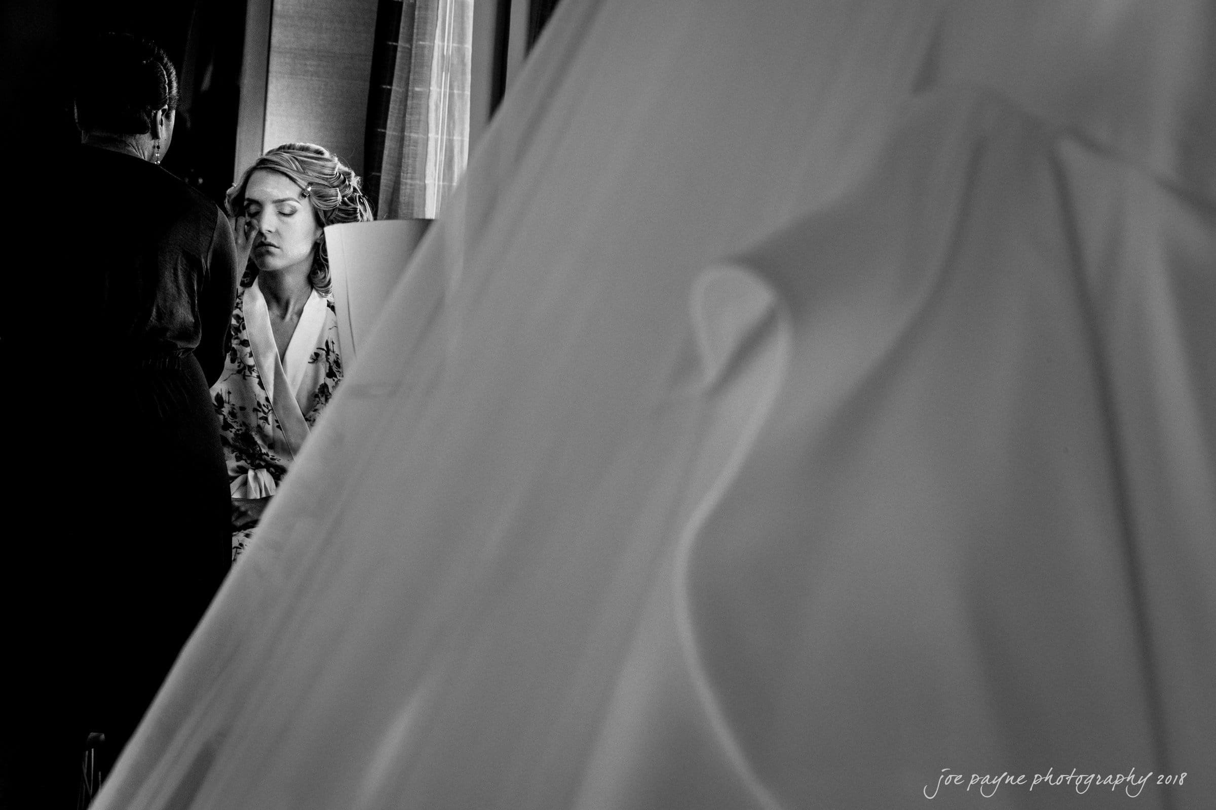 Black and white image of a bride getting ready, with eyes closed, as a makeup artist applies makeup. The foreground features a blurred, draped wedding dress, adding depth to the scene. The atmosphere is calm and focused.
