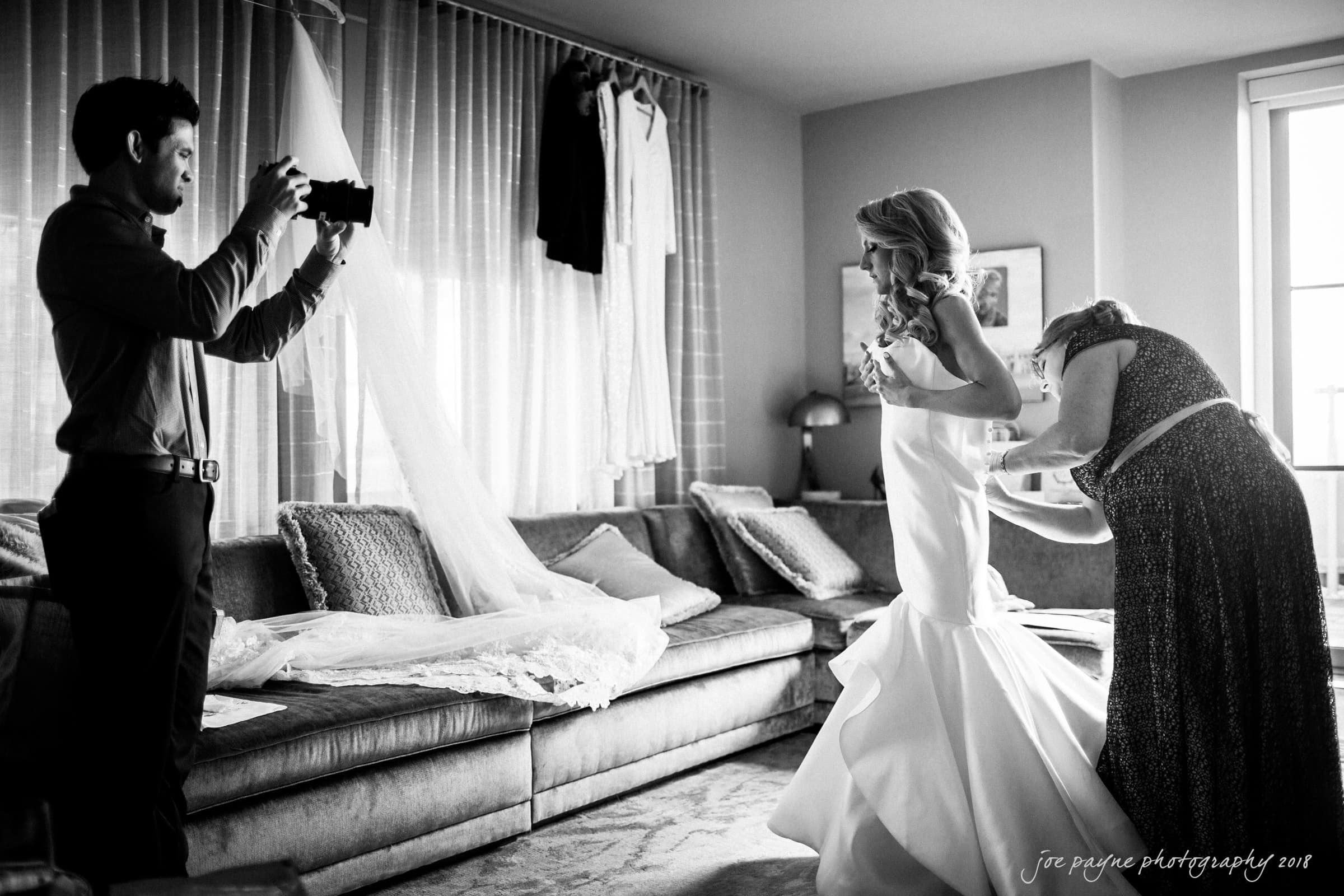A bride in a white gown is being helped by a woman, likely the mother, with her dress in a well-lit room. A photographer captures the moment. A wedding dress train is draped over a couch in the background. The setting is elegant and intimate.