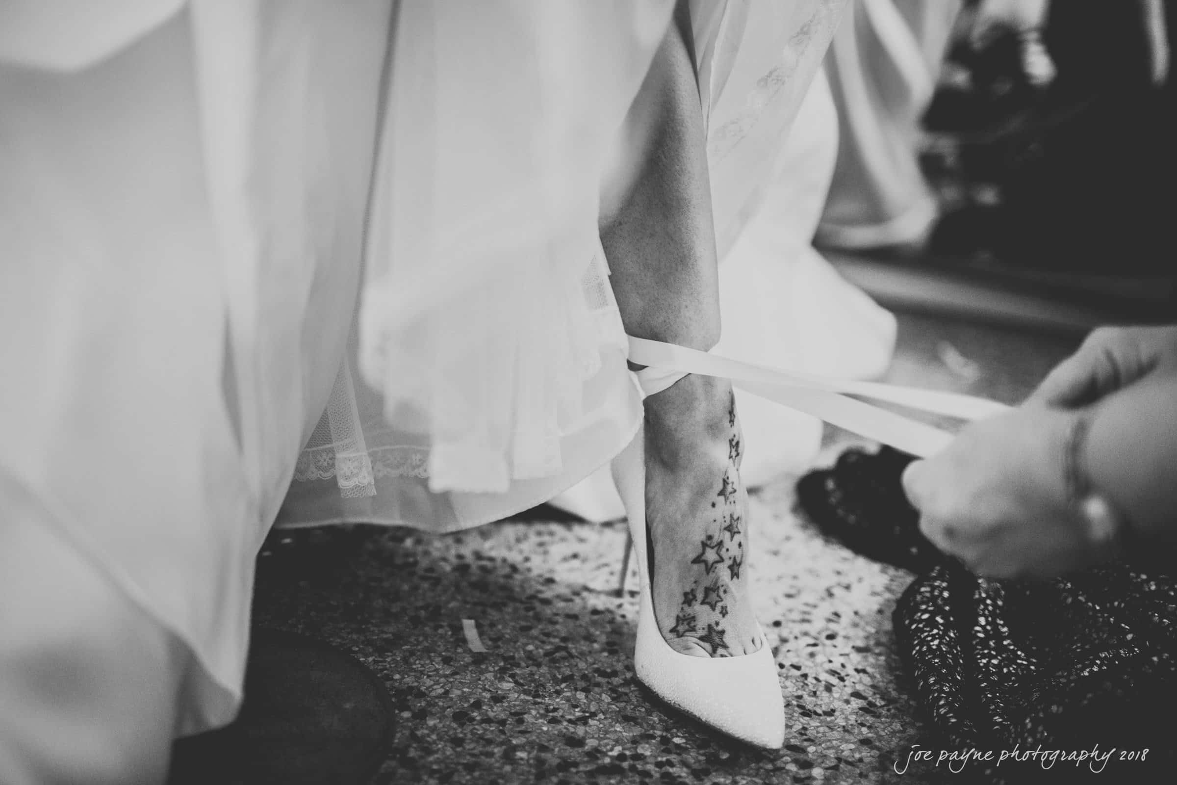 A black and white photo of a brides foot being assisted into a white high heel. The brides ankle has a star tattoo. The heel features lace ankle straps being secured by another person. The wedding dress fabric is partially visible.