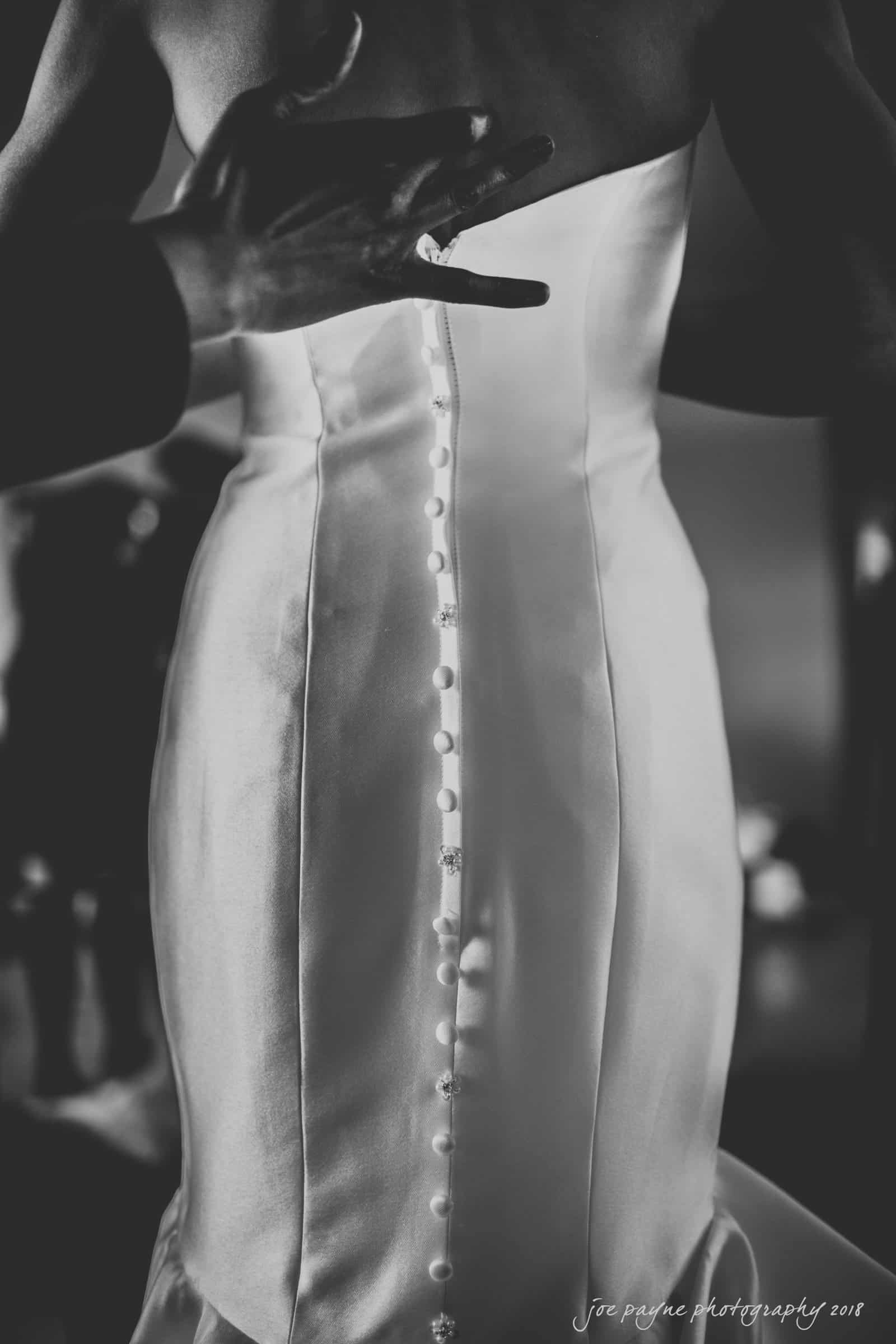 A black and white photo of a person fastening buttons on the back of a satin wedding dress. The focus is on the row of buttons running down the back. Hands are visible, adjusting the dress.