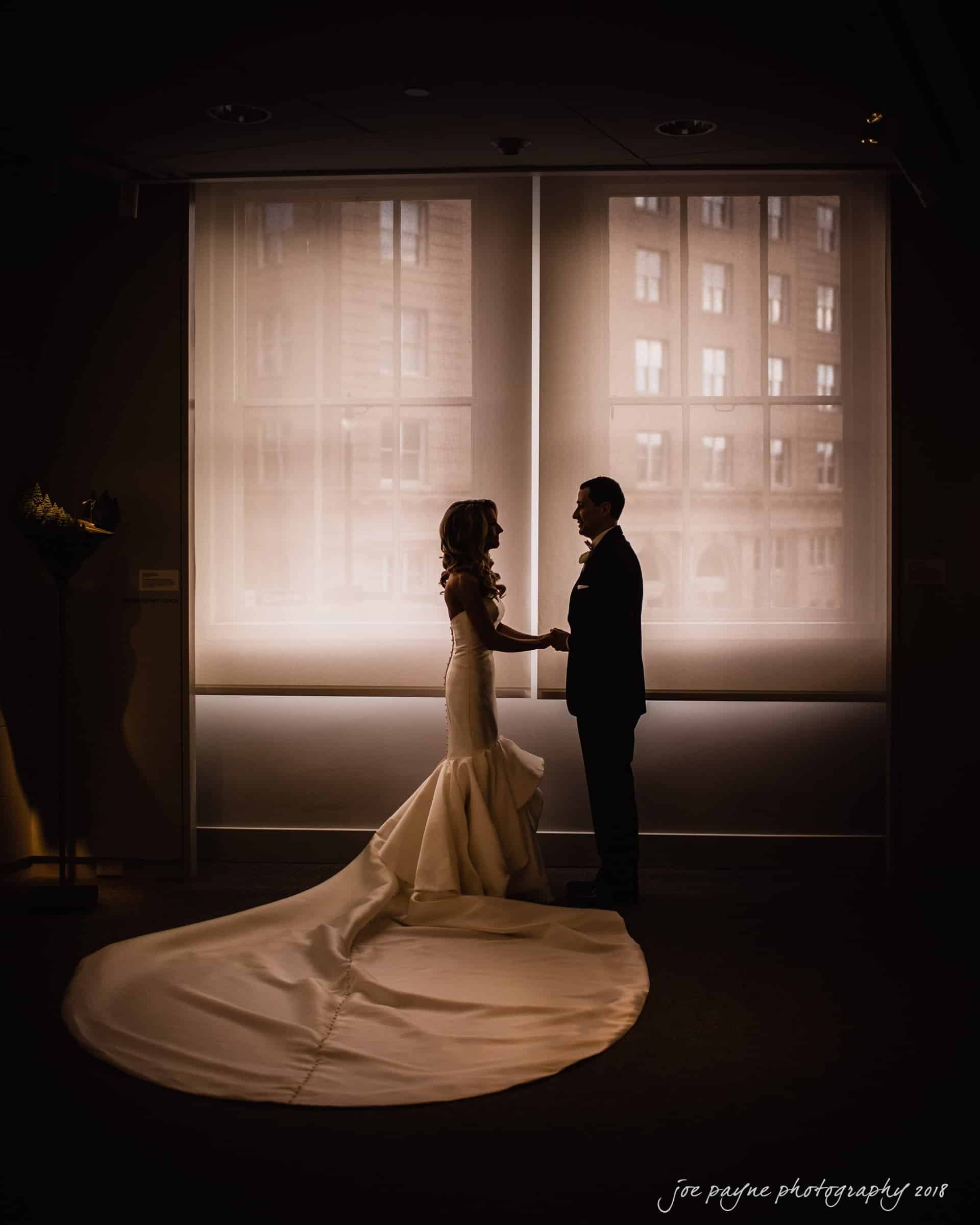 A bride and groom stand facing each other in front of large, softly lit windows. The bride wears a long, flowing gown with an extended train, while the groom is in a dark suit. Their silhouettes are highlighted against the bright backdrop.