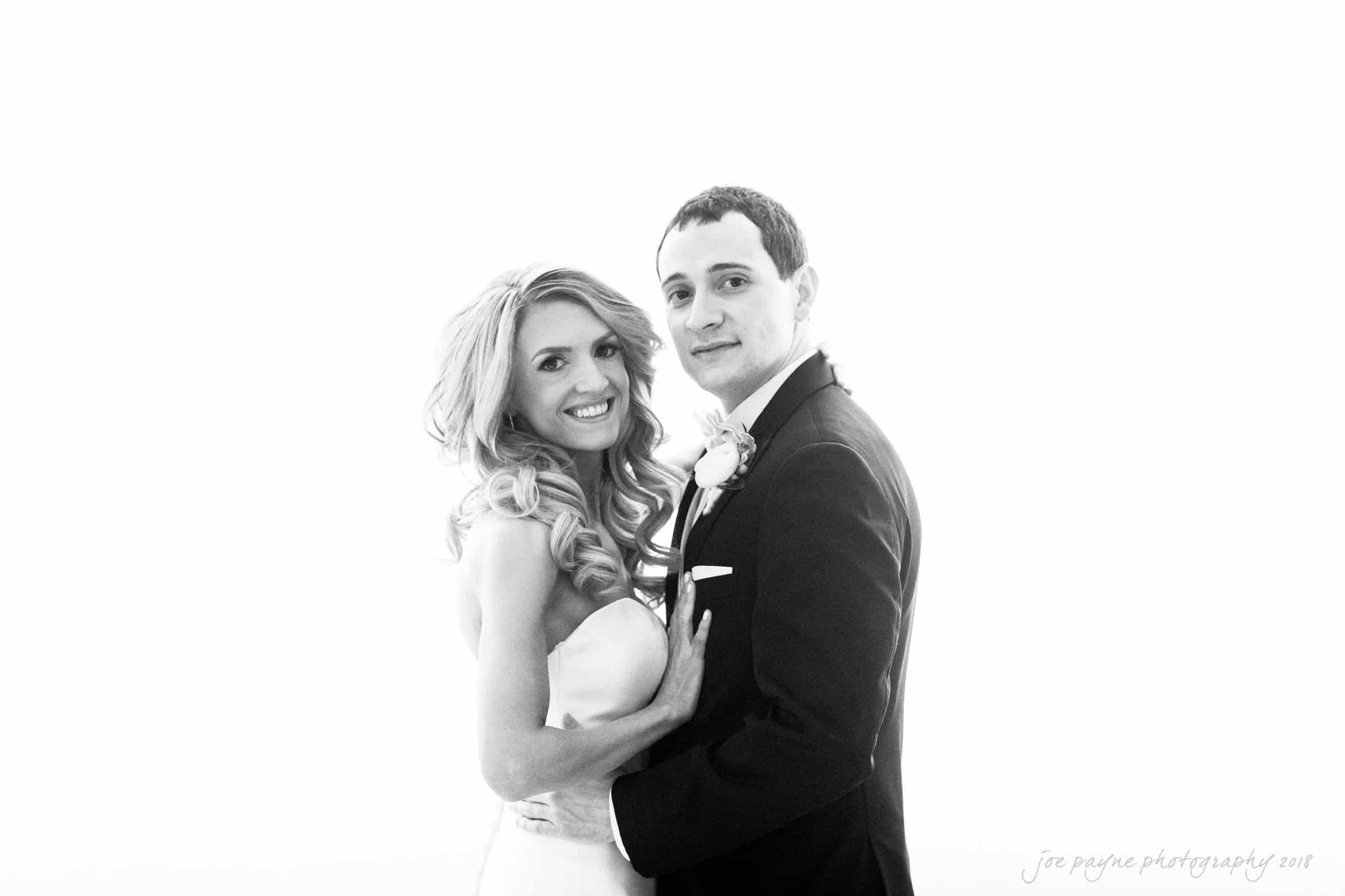 A black and white photo of a couple on their wedding day. The bride is wearing a strapless gown with long, curly hair, and the groom is in a suit with a boutonniere, standing together and smiling at the camera.