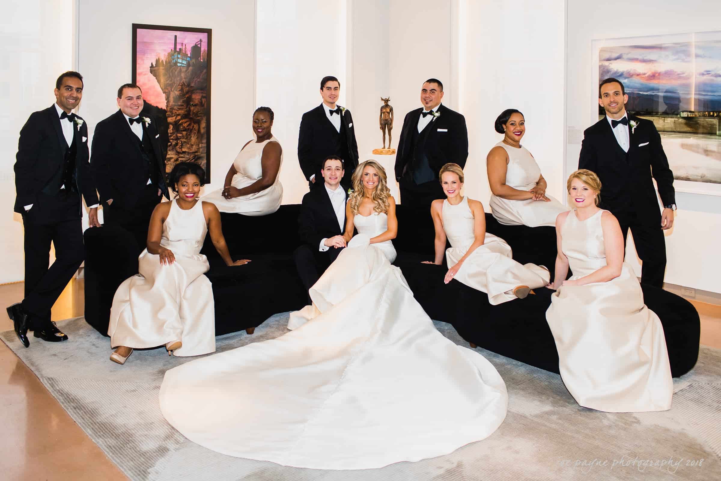A wedding party poses elegantly indoors against a backdrop of modern art. The bride is seated at the center in a white gown with a long train, surrounded by bridesmaids in cream dresses and groomsmen in black suits. There are smiles and relaxed poses.