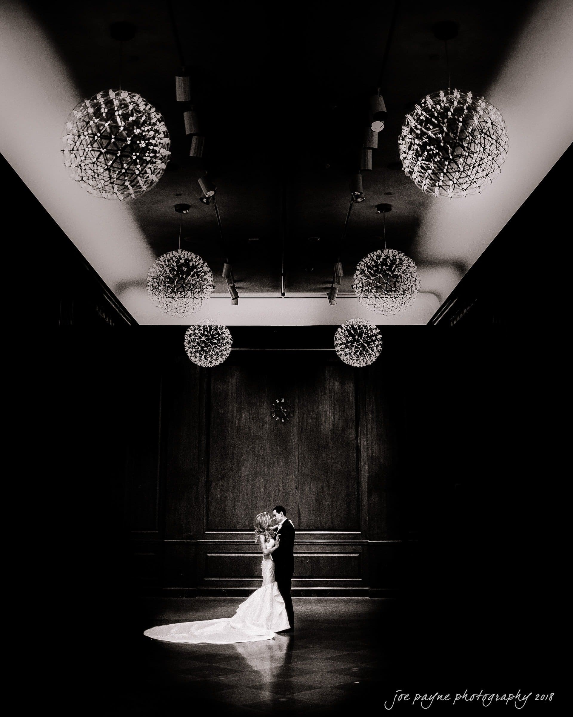 A newlywed couple shares a first dance in a dimly lit, elegant ballroom. The brides flowing gown trails behind her. Spherical chandeliers hang from the ceiling, casting soft light on the scene.