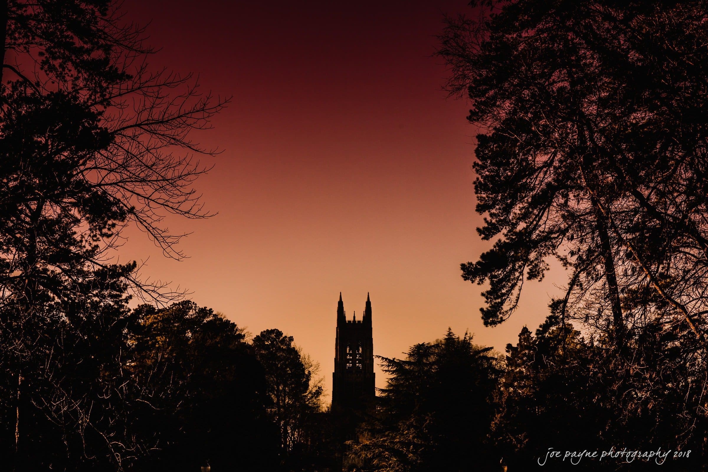Silhouette of a church tower at sunset, surrounded by trees on either side. The sky transitions from deep orange to dark red, creating a dramatic backdrop.