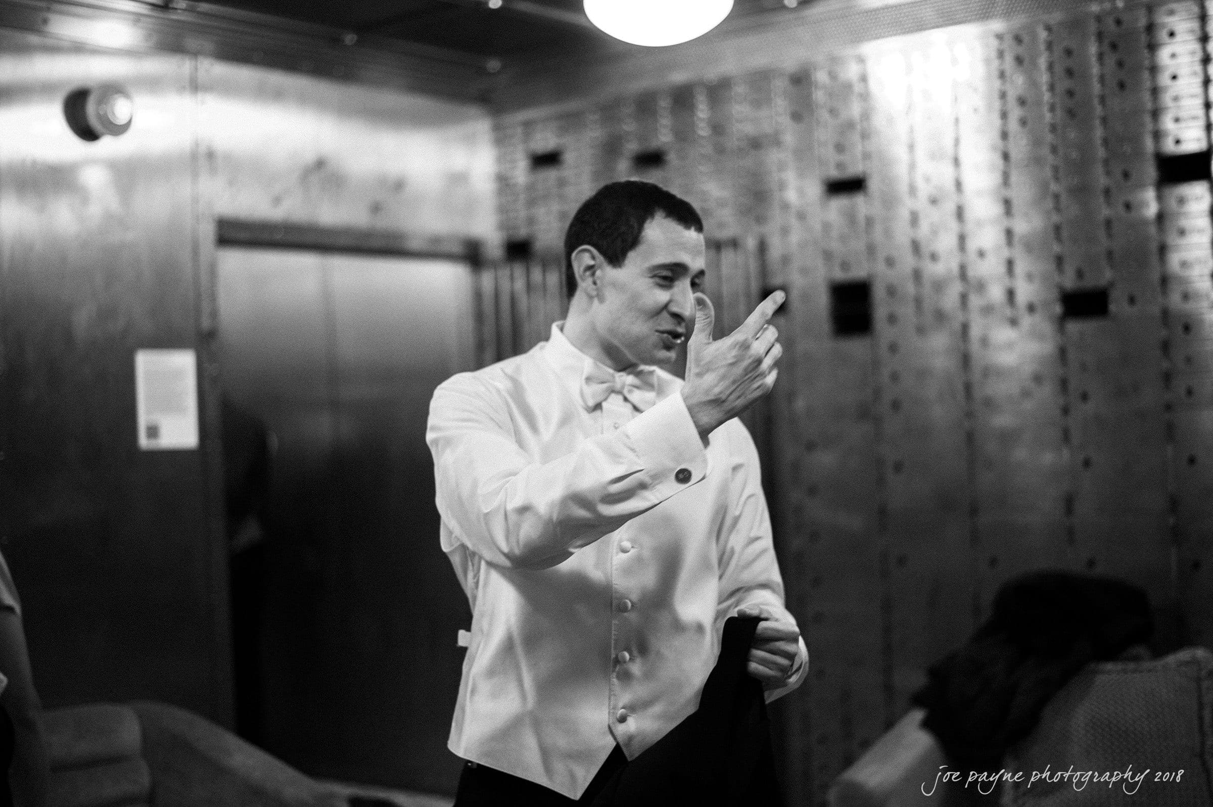 A man in a formal white shirt and bow tie points while holding a jacket. The setting is an elegant room with metallic walls and a vintage feel. The black and white image has a candid, lively atmosphere.
