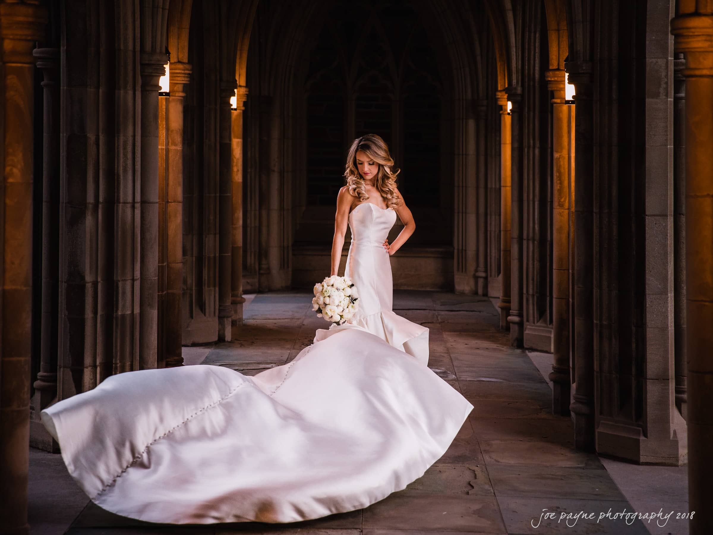 A bride in a flowing white gown stands in a dimly lit corridor with stone arches. She holds a bouquet of white flowers and gazes downward, with her train elegantly spread behind her on the stone floor.