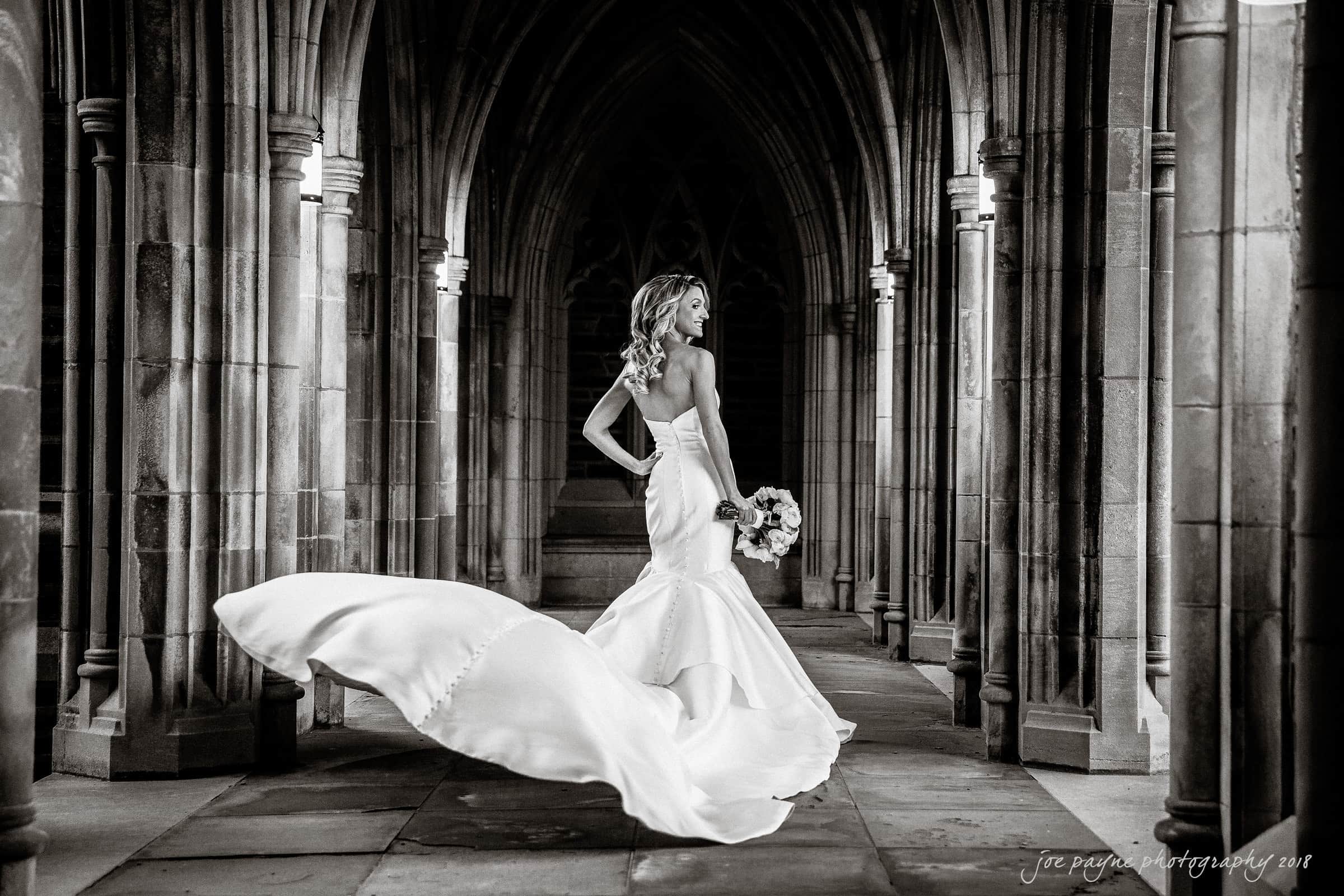 A bride in a flowing wedding dress walks gracefully down a dimly lit, Gothic-style stone corridor. She holds a bouquet in one hand, and the train of her dress billows elegantly behind her. The scene is black and white, creating a timeless feel.