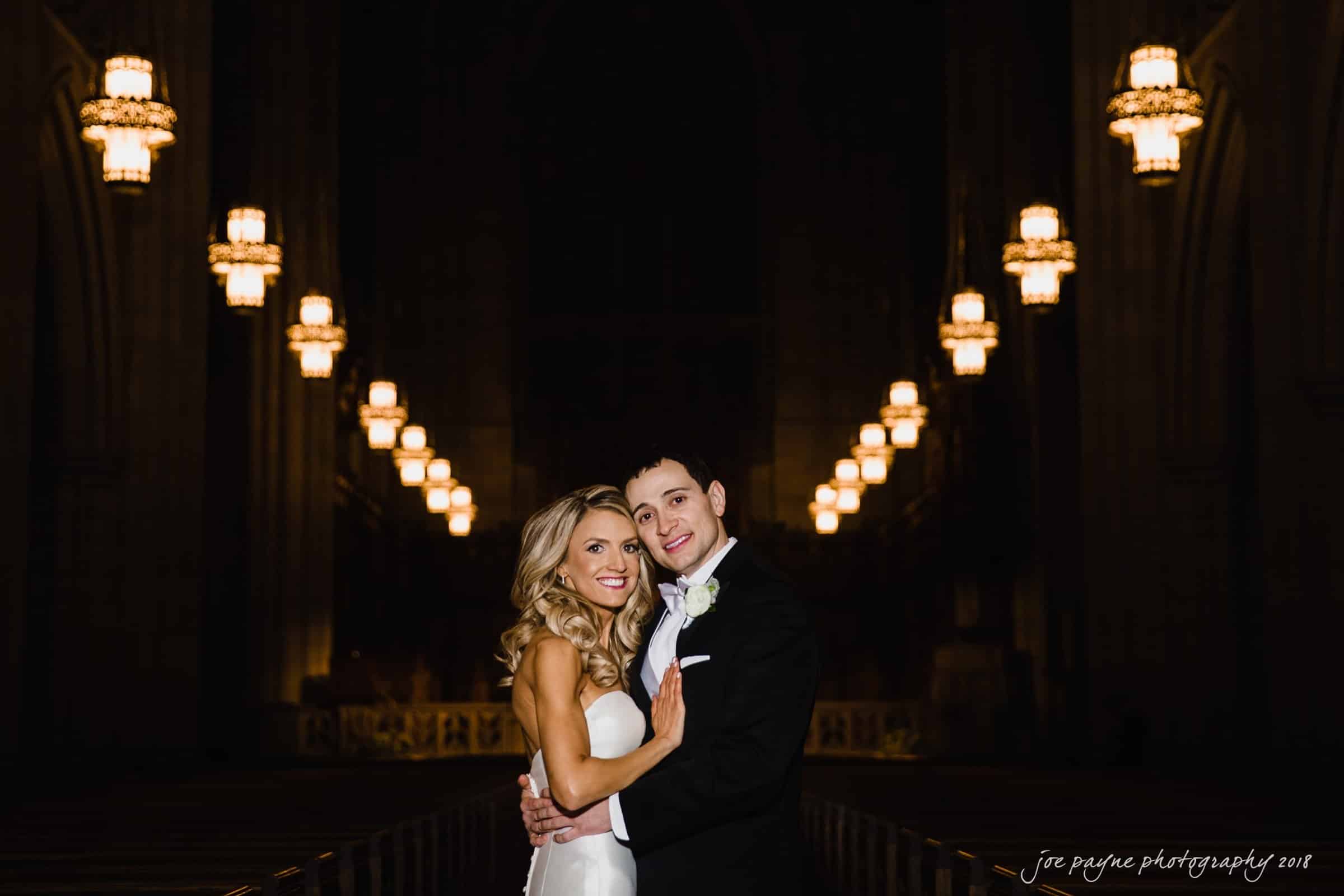 A bride and groom pose smiling in an ornately lit cathedral. The bride wears a white dress and the groom is in a black suit with a white boutonniere. Hanging lights lead toward the altar in the background.