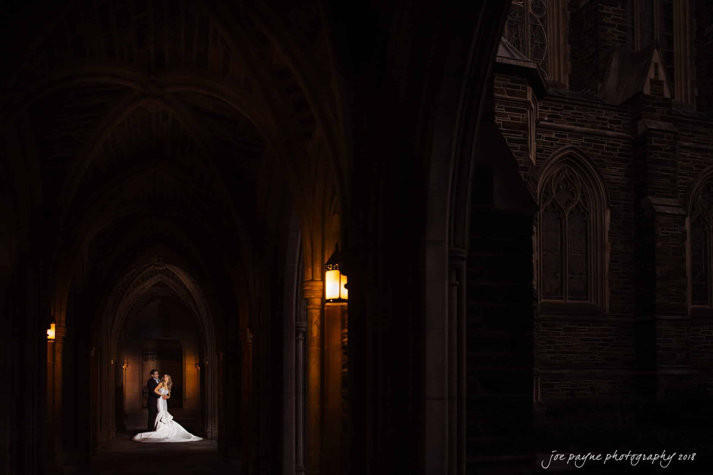 A bride and groom stand close together in a dimly lit stone archway. Warm light illuminates them, creating a dramatic contrast against the dark, gothic-style architecture.