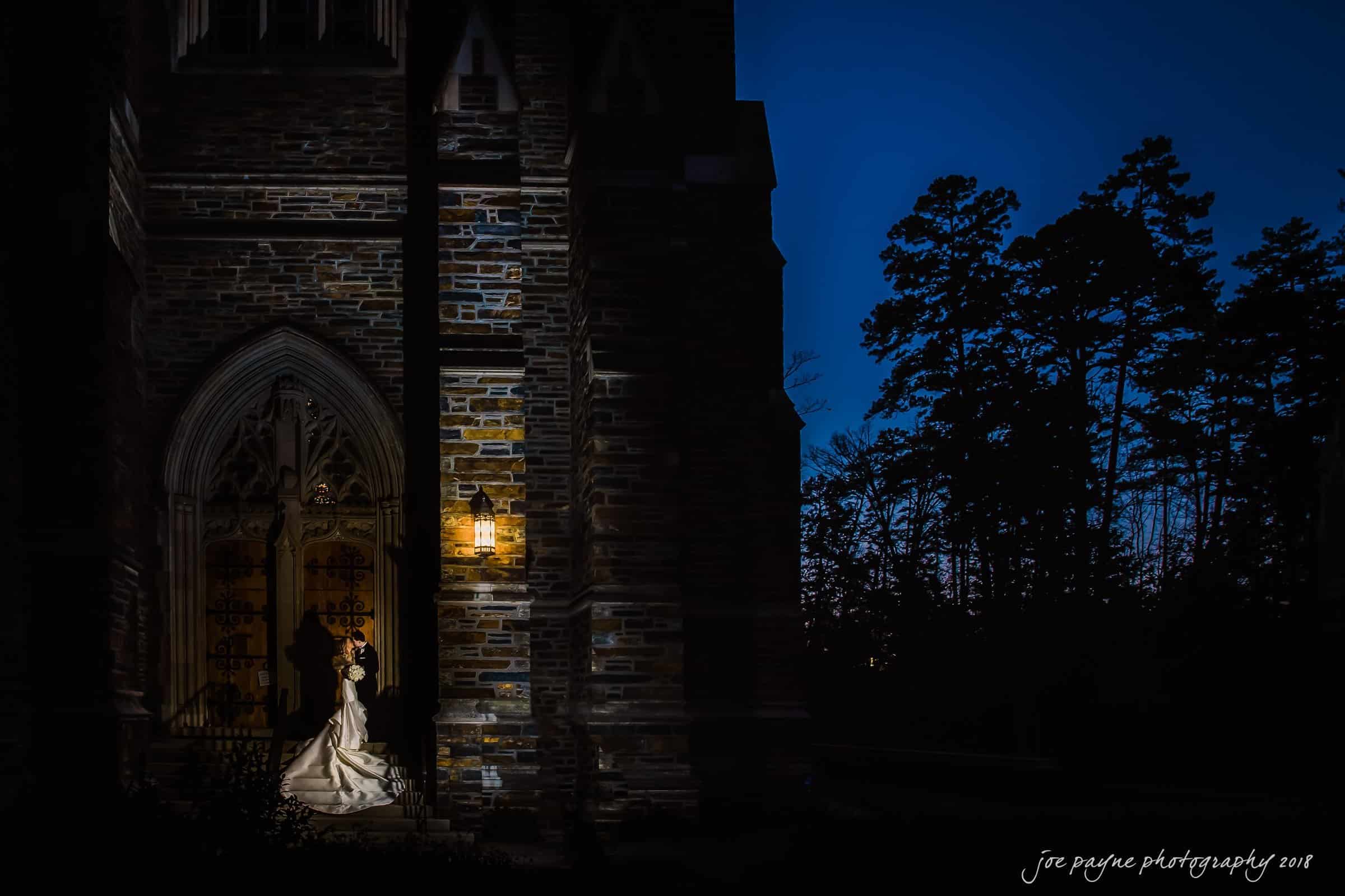 A bride and groom share a romantic moment under a glowing lamp by a stone archway of a church at night. The scene is set against a dark, clear sky with silhouettes of tall trees in the background.