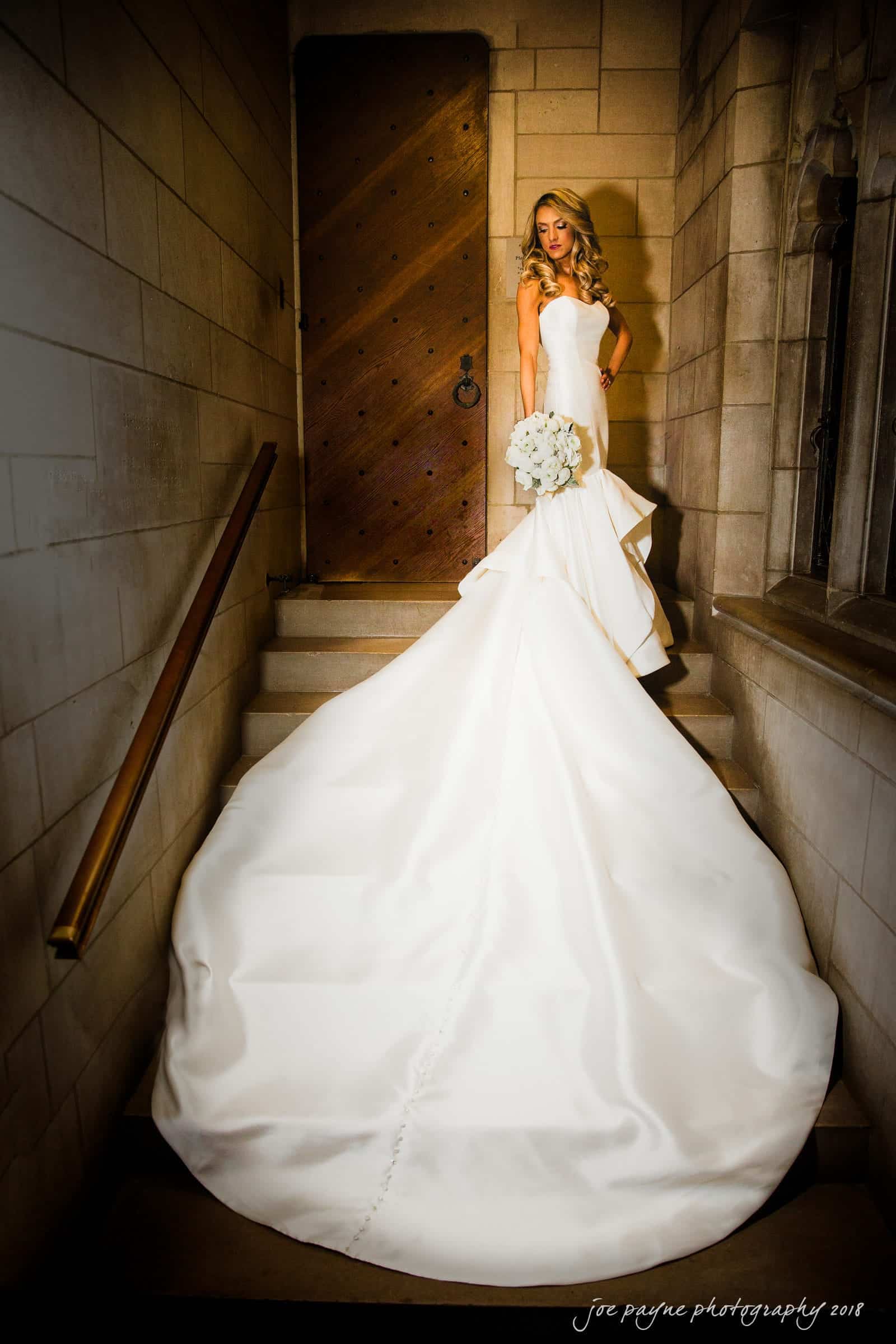 A bride in a strapless wedding gown stands on stairs, holding a white bouquet. The gown has a long flowing train, cascading down the steps. She is positioned beside a wooden door in a stone-walled setting.