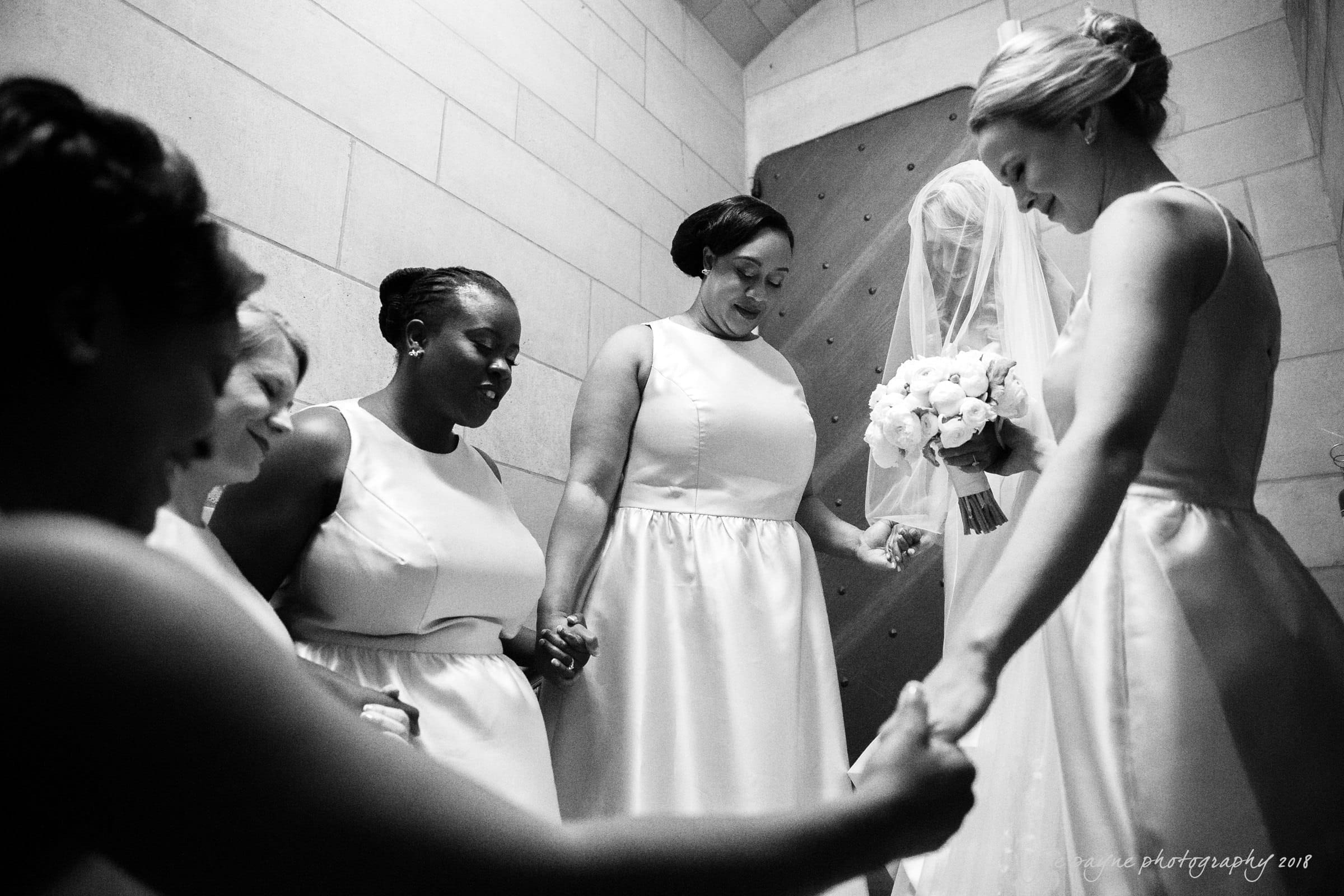 A group of women in elegant dresses gather closely in a circle, holding hands and bowing their heads, appearing to share a moment of reflection or prayer. A bride stands among them, holding a bouquet of flowers. The setting is a softly lit room.