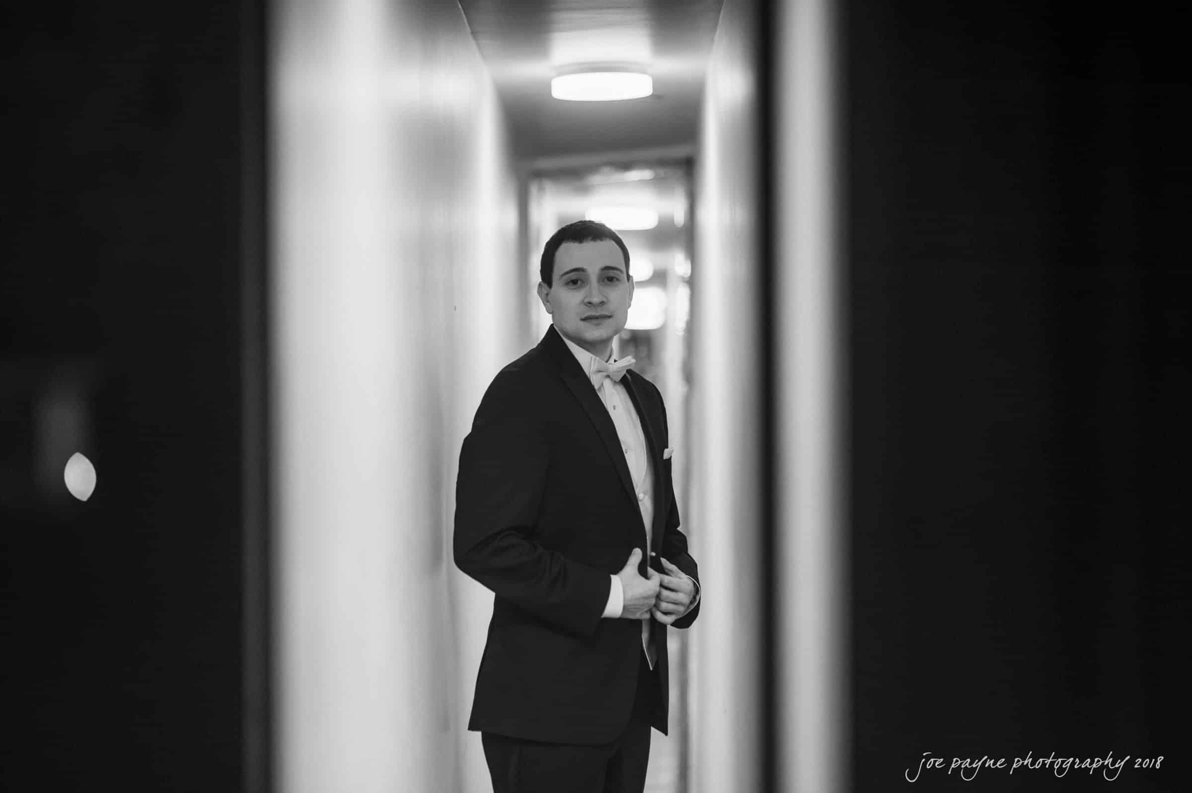 A man in a tuxedo stands in a narrow, well-lit hallway, adjusting his jacket and looking towards the camera. The background is softly blurred, highlighting his formal attire.