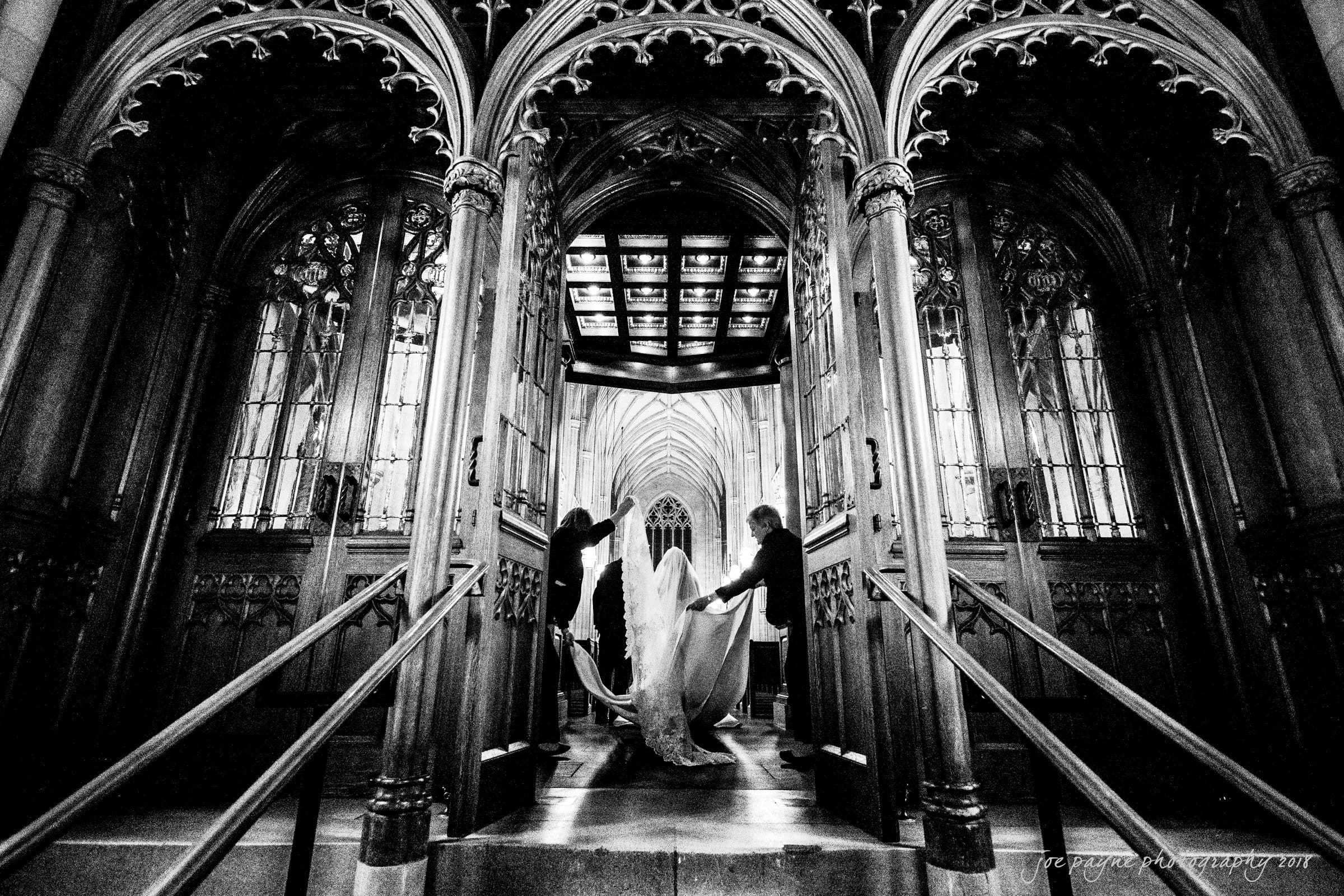 A bride in a flowing gown is assisted by two people outside a grand cathedral entrance with ornate wooden doors. The interior is illuminated, highlighting the intricate arches and Gothic architecture. Black and white image.